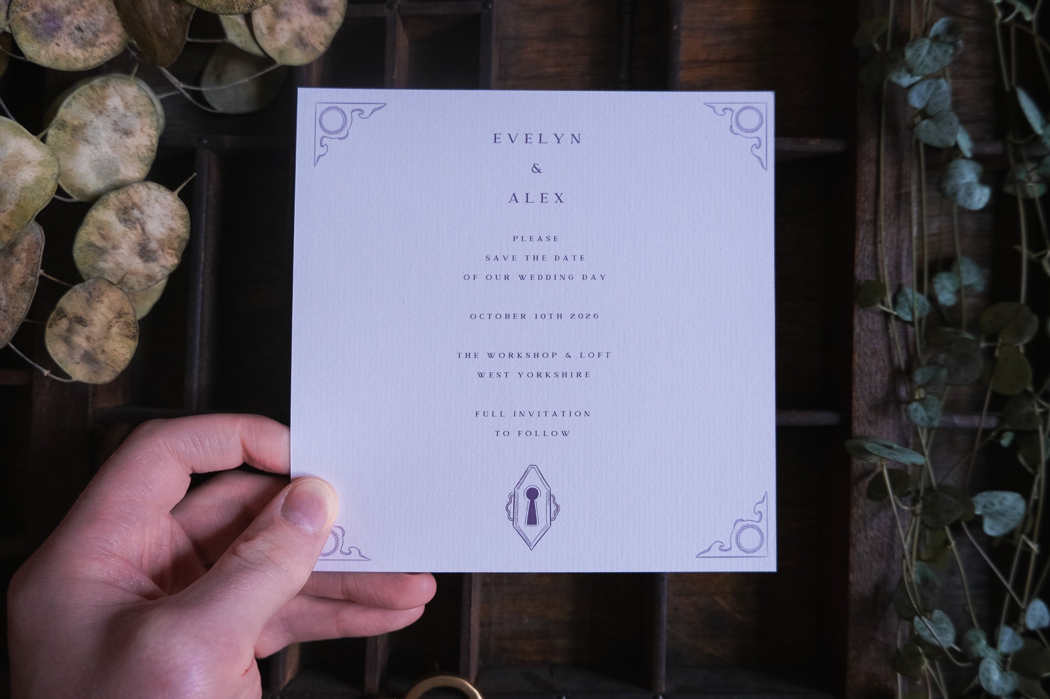 A hand holds a luxury vintage Save the Date card with heart padlock and skeleton key motifs on rustic wood with dried leaves.