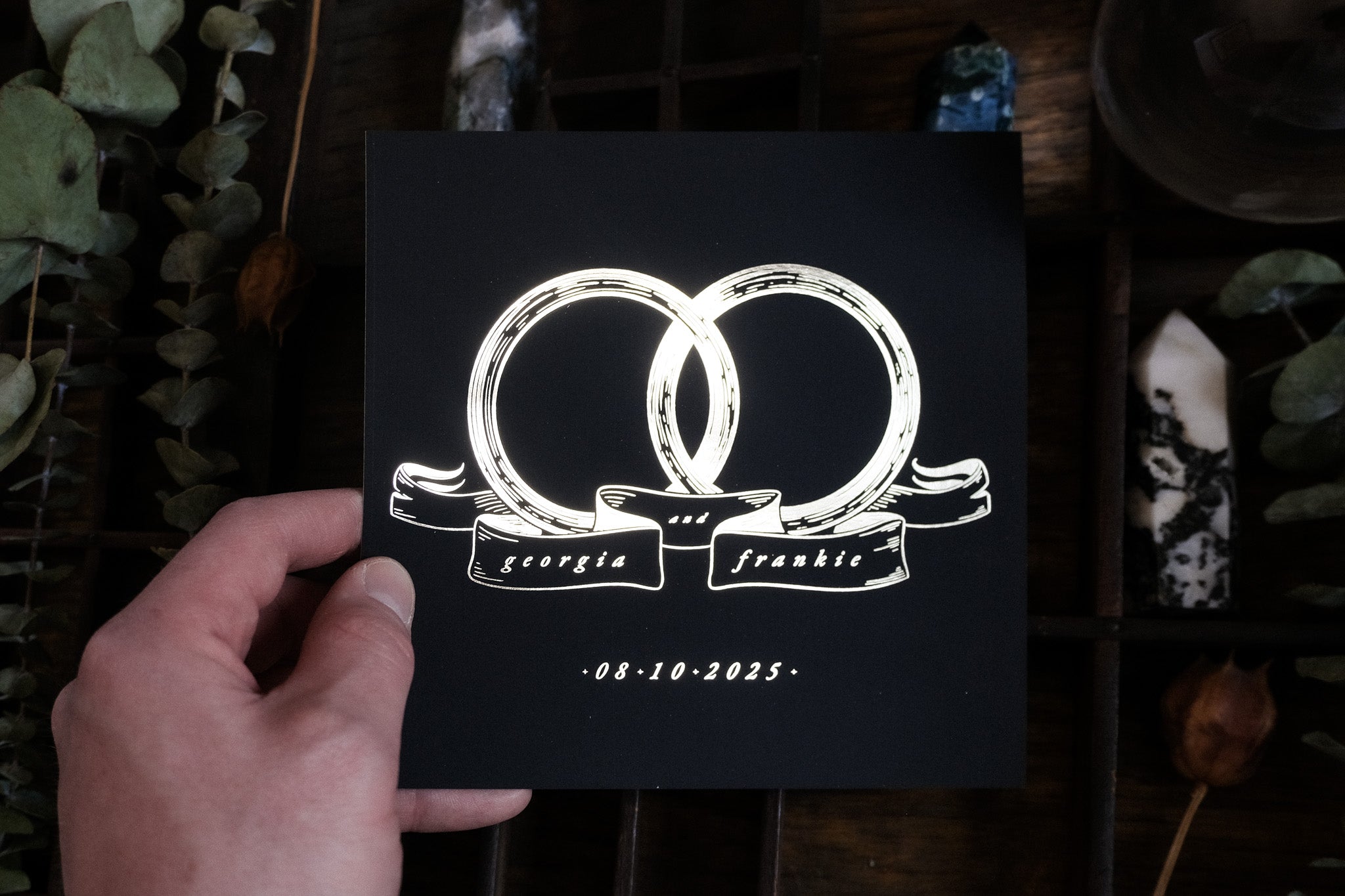 A hand displays luxury UK wedding stationery with interlocking rings and gold foil, set amid greenery and crystals for a romantic touch.