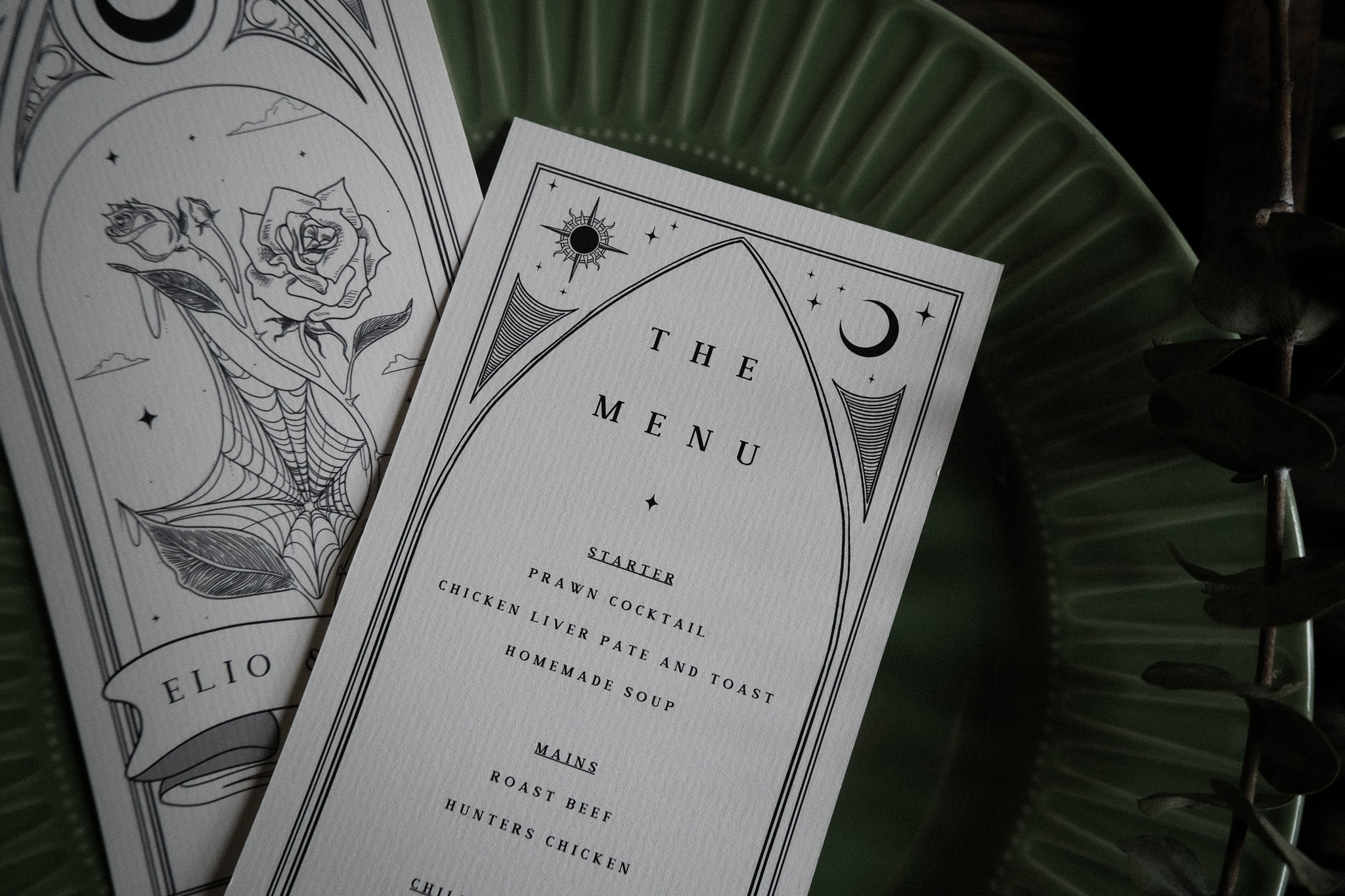 Close-up of luxury UK wedding menu cards with gothic romantic design, monochrome rose and spiderweb art, elegant type, dramatic appeal.