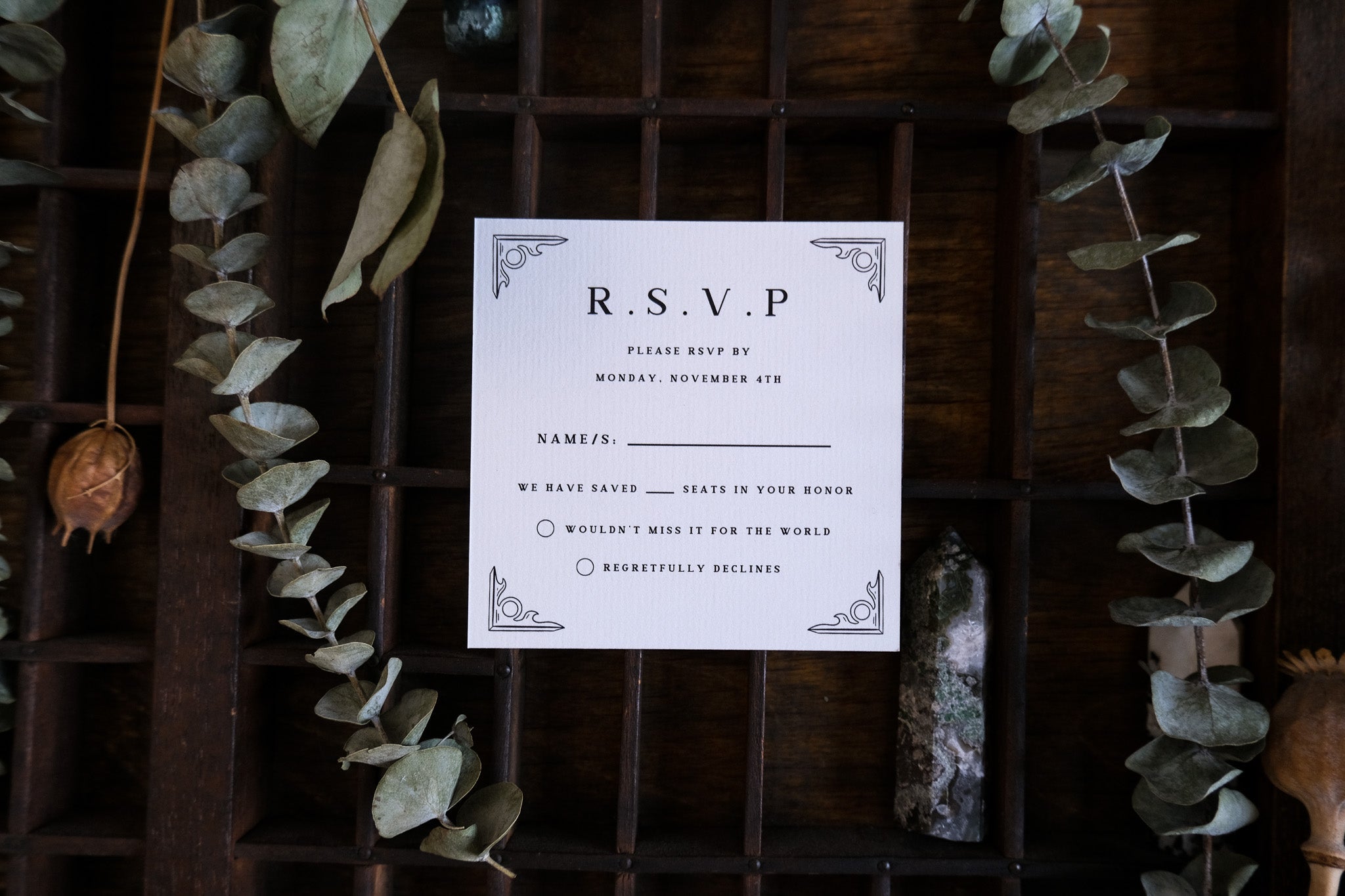 Luxury UK wedding RSVP card featuring vintage botanical motifs, dramatic decorative corners, and boho dried leaves on rustic wood.