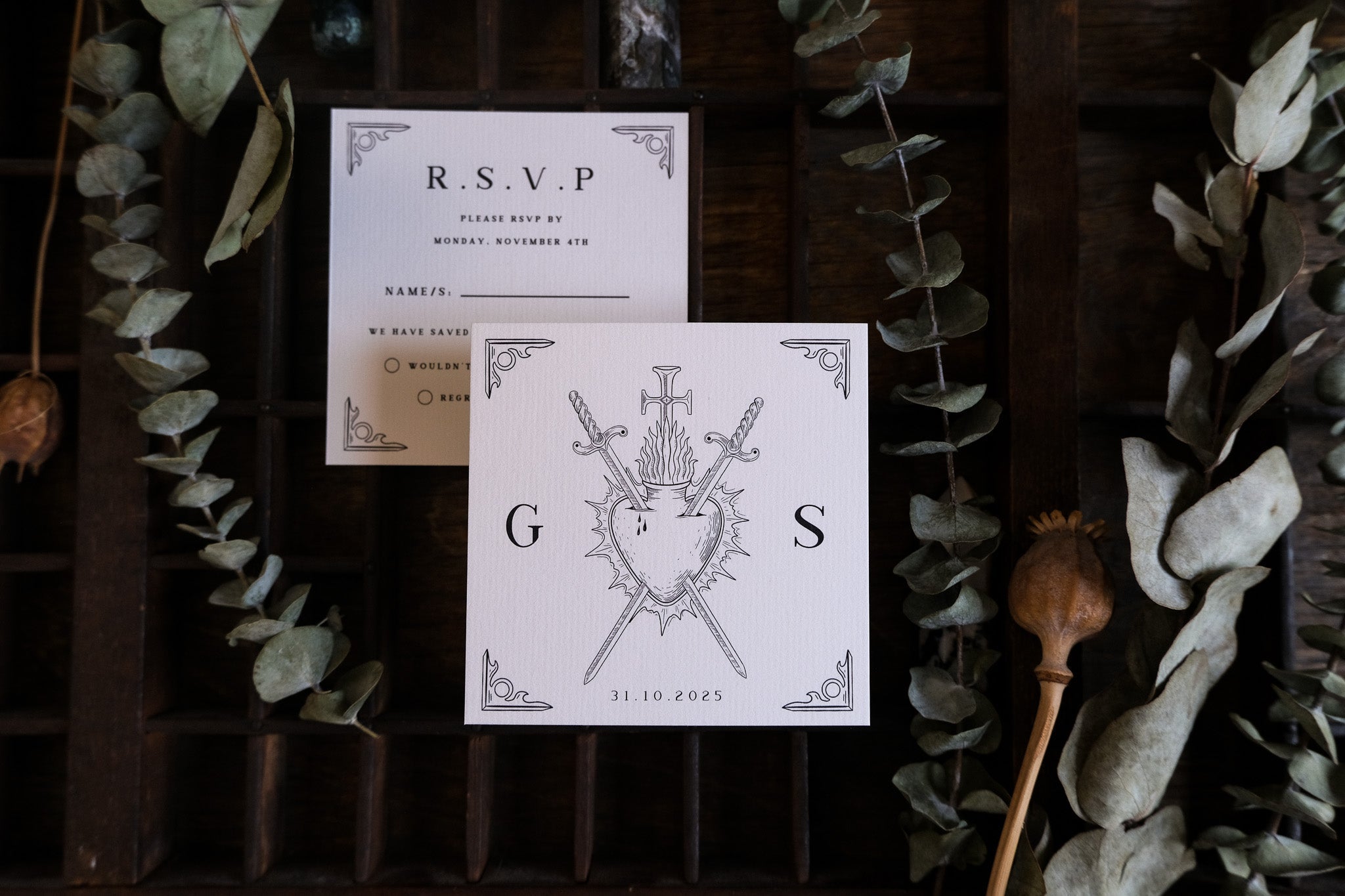Luxury gothic wedding RSVP card with sword and shield motifs on premium card, displayed on dark wood with dried eucalyptus accents.