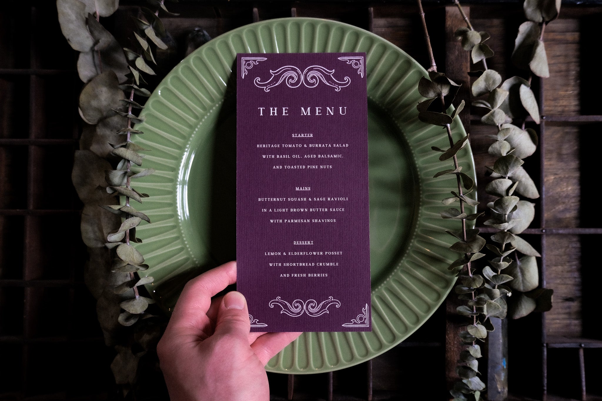 A hand displays a luxury tarot-inspired wedding menu card with ornate vintage borders, on a green plate amid dried leaves.
