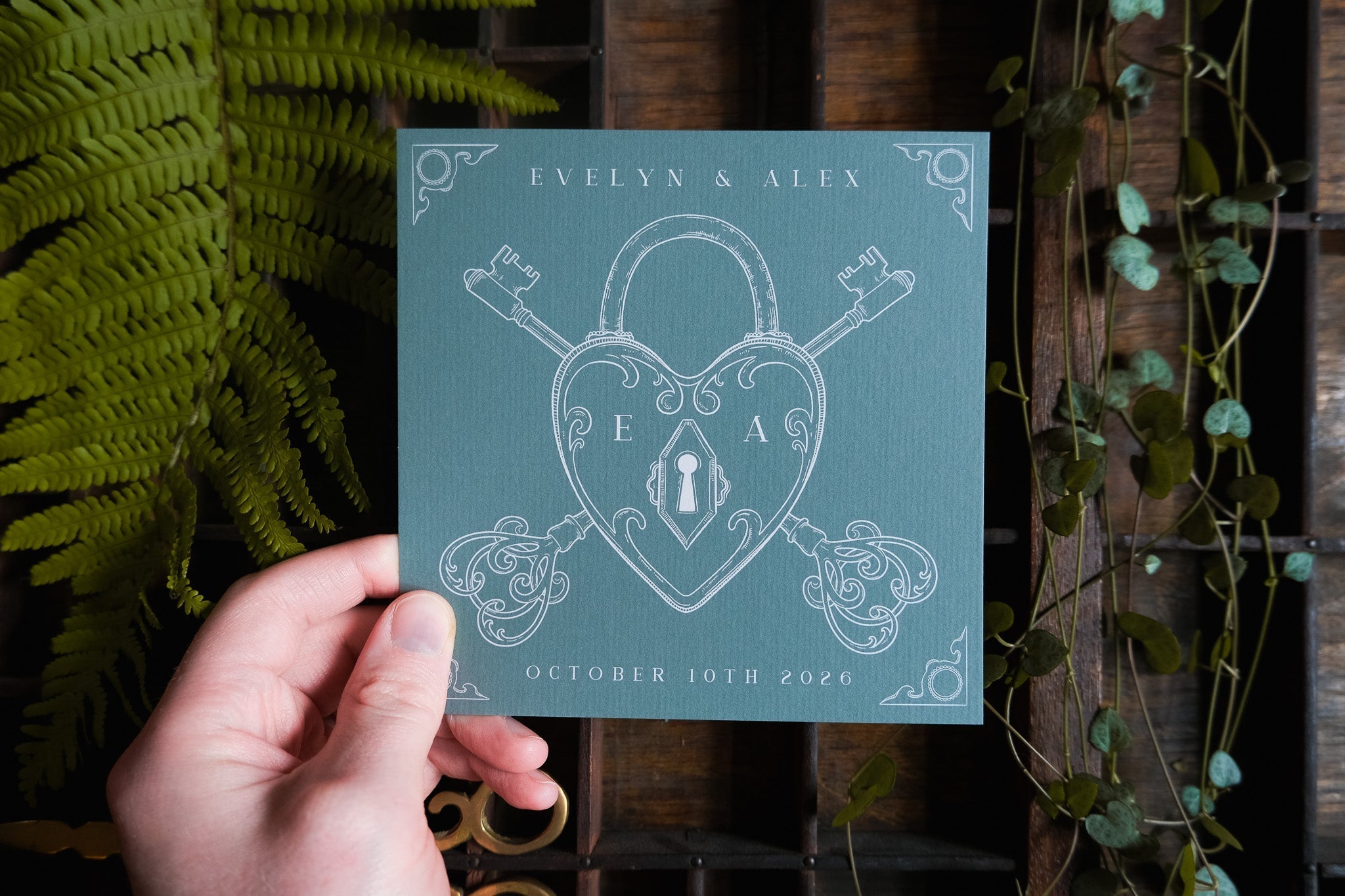 A hand presents a luxury vintage-style Save the Date card in teal, featuring gothic lock and key motifs amid lush botanical accents.
