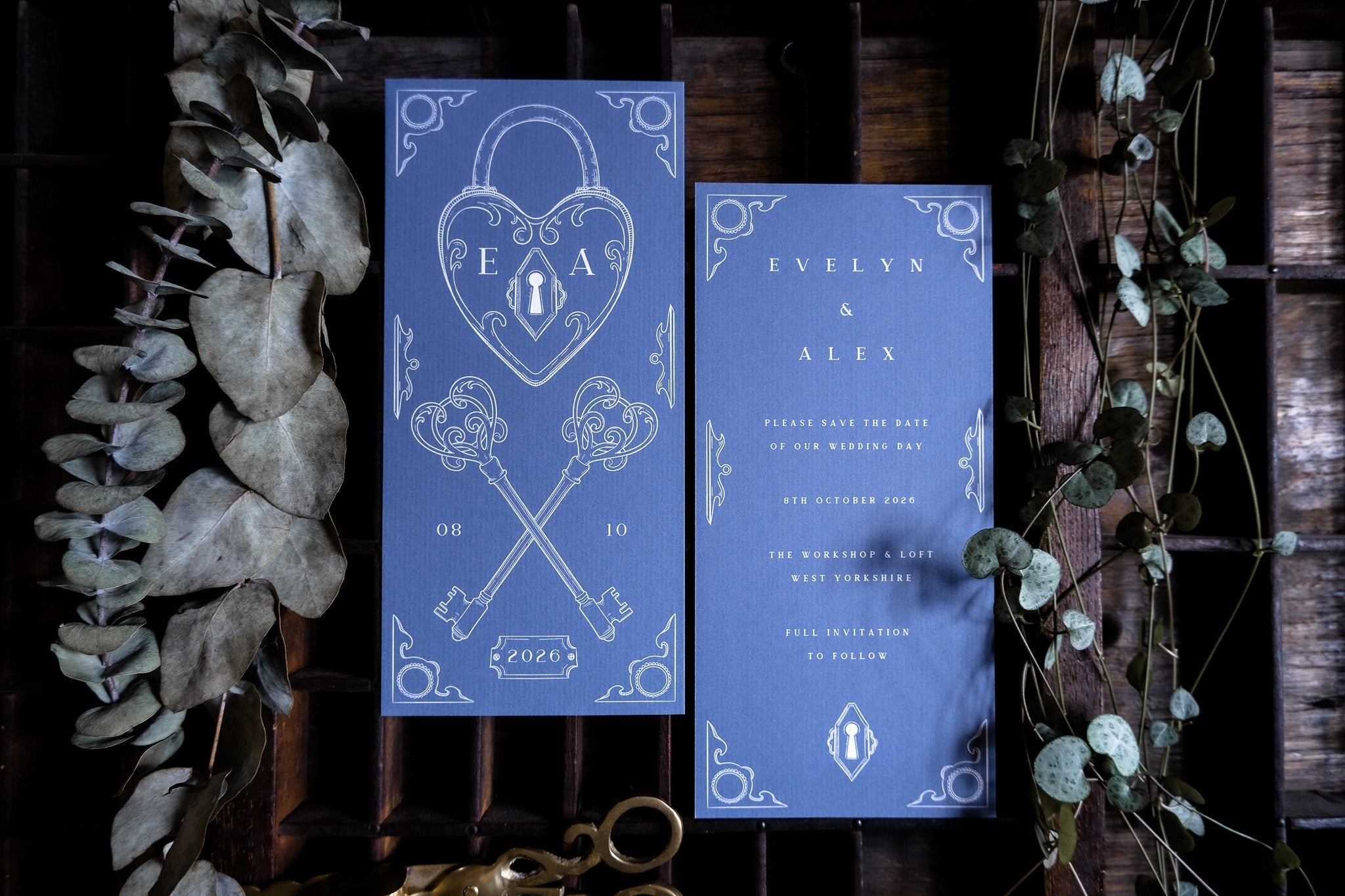 Luxury vintage-inspired save the date cards in deep blue with ornate Victorian borders, keys and heart locks, styled amid dried leaves.