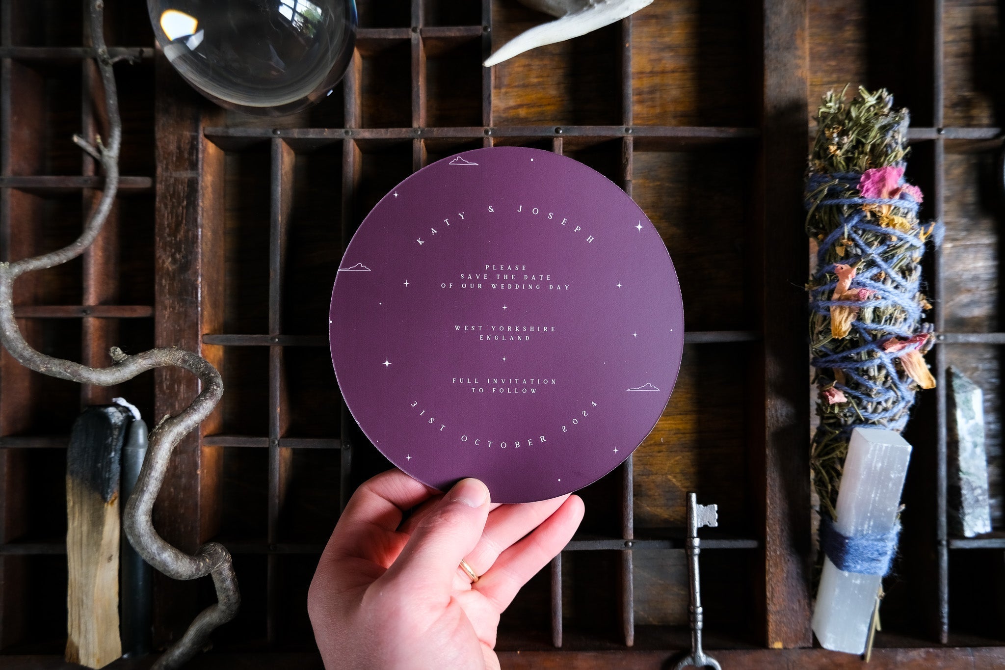 A hand holds a luxury gothic circular wedding stationery card with white text, set on a tray with crystals, dried herbs, and vintage accents.