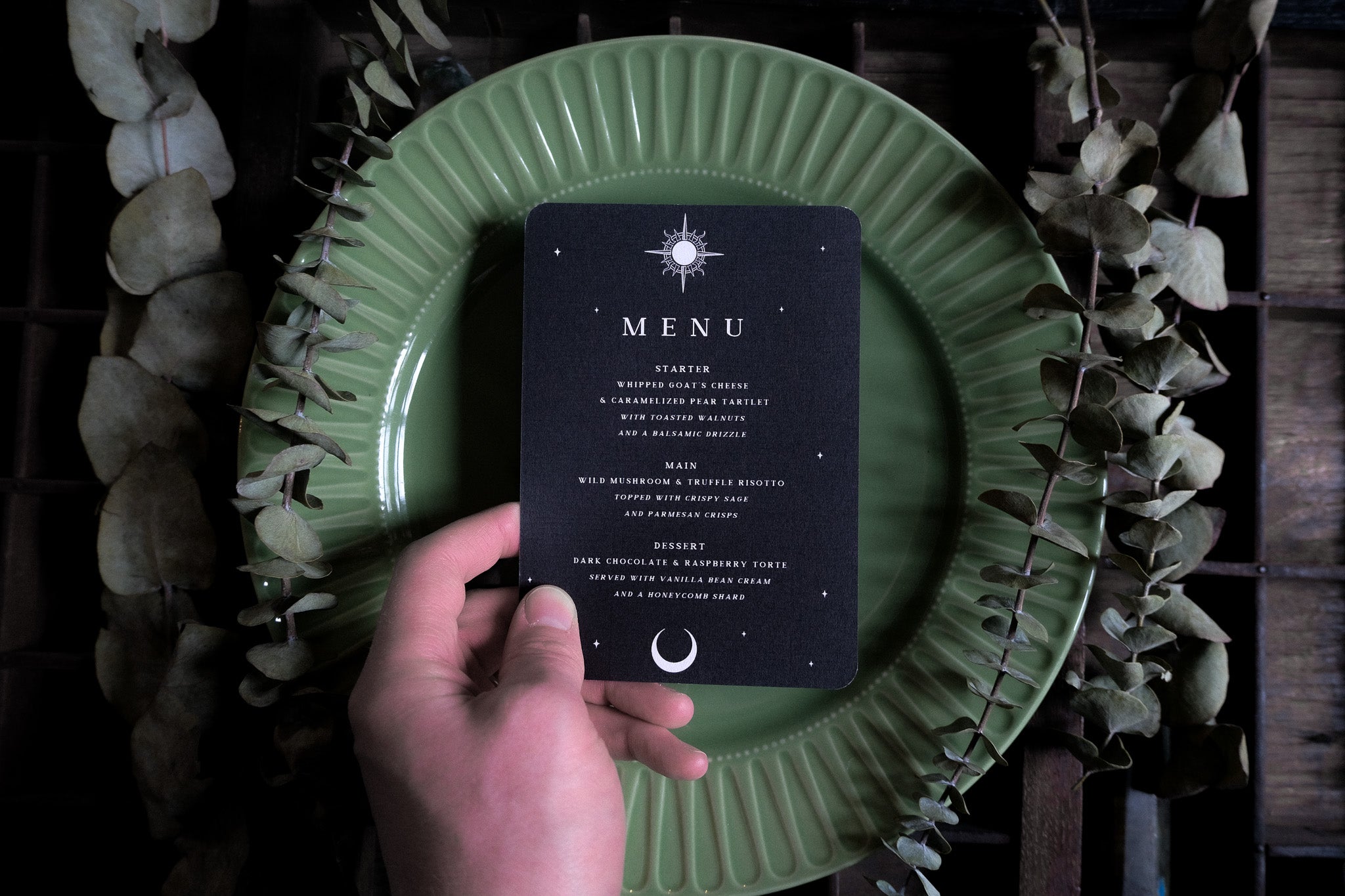 A hand holds a luxury gothic wedding menu card with a skeleton couple design, displayed on a ribbed green plate with dried leaves.