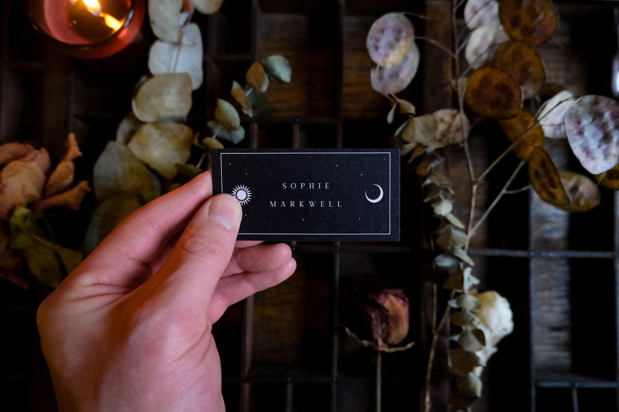 A hand holds a luxury black wedding placecard with gold foil celestial sun and moon design, styled amid dried leaves and candlelight.