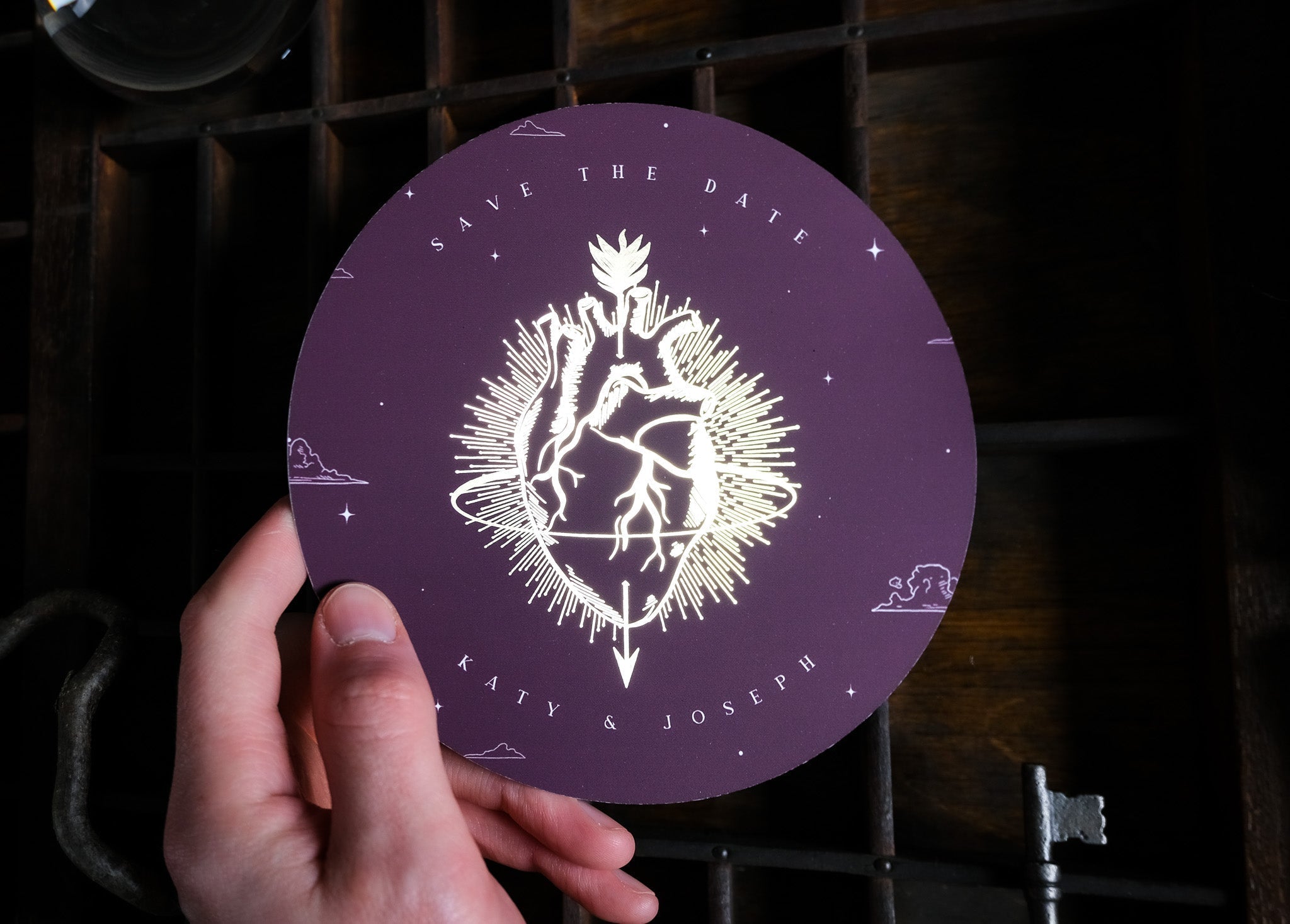 A hand displays a luxury circular save the date card with gold foiling, showcasing a gothic romantic anatomical heart design.