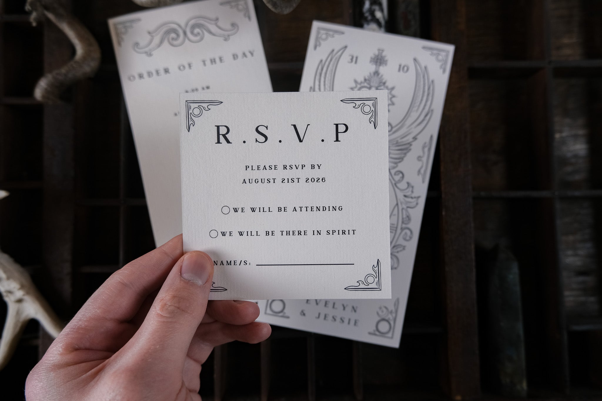 A hand holds a square RSVP card; luxury gothic wedding stationery with dramatic, vintage-inspired details rests on dark wood.