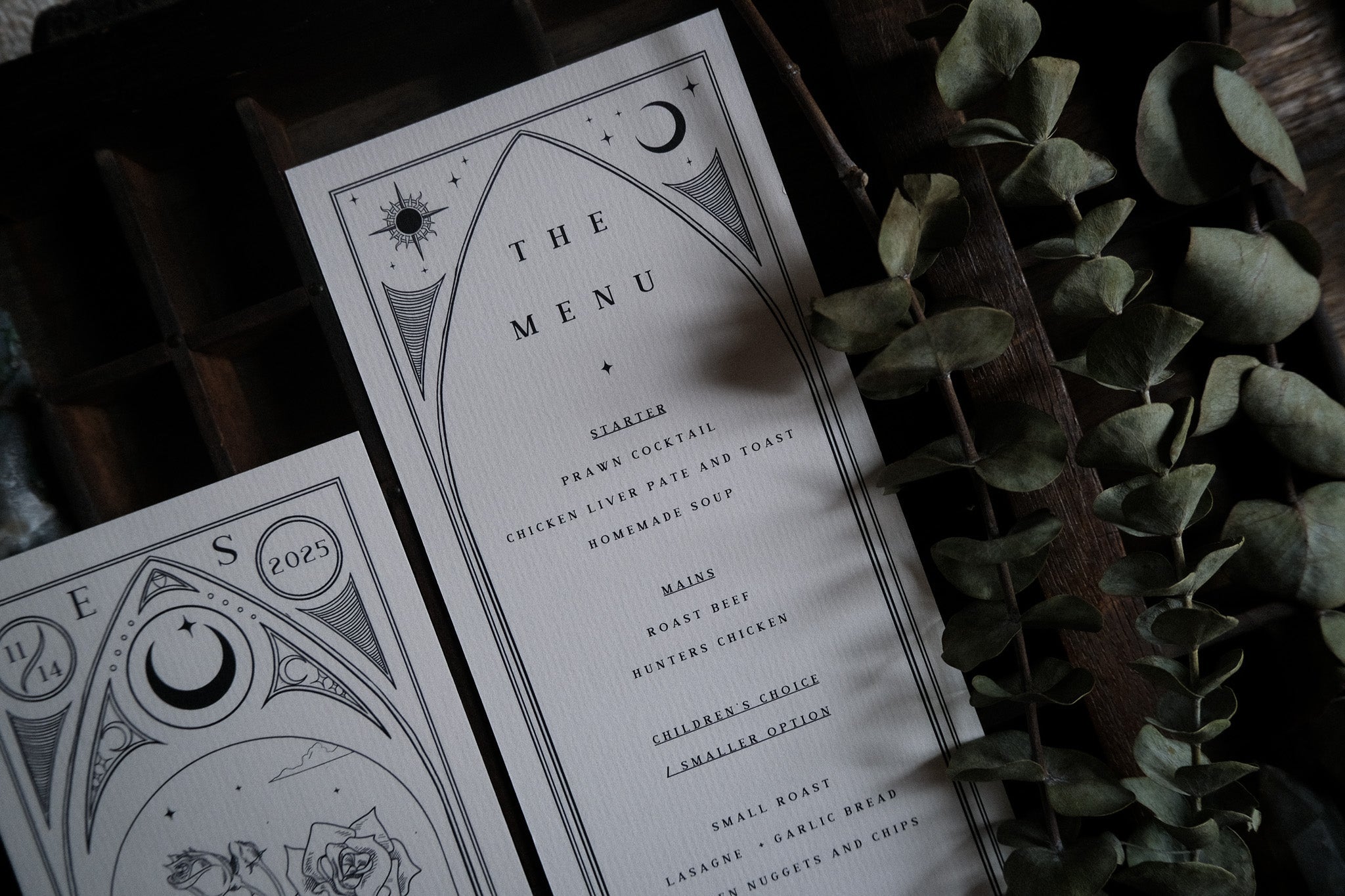 Luxury UK wedding menu cards in a gothic, romantic style with celestial and floral motifs, set on wood beside dried eucalyptus.