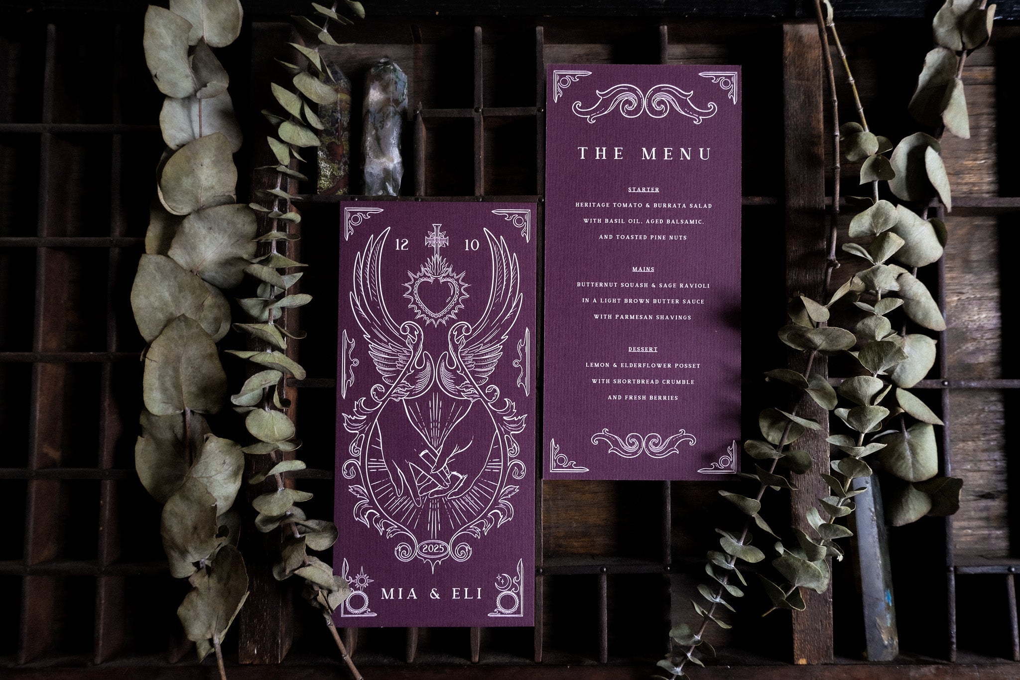 Luxury gothic tarot-inspired wedding menu cards in deep purple with ornate white detailing and bird motifs, styled with eucalyptus on dark wood.
