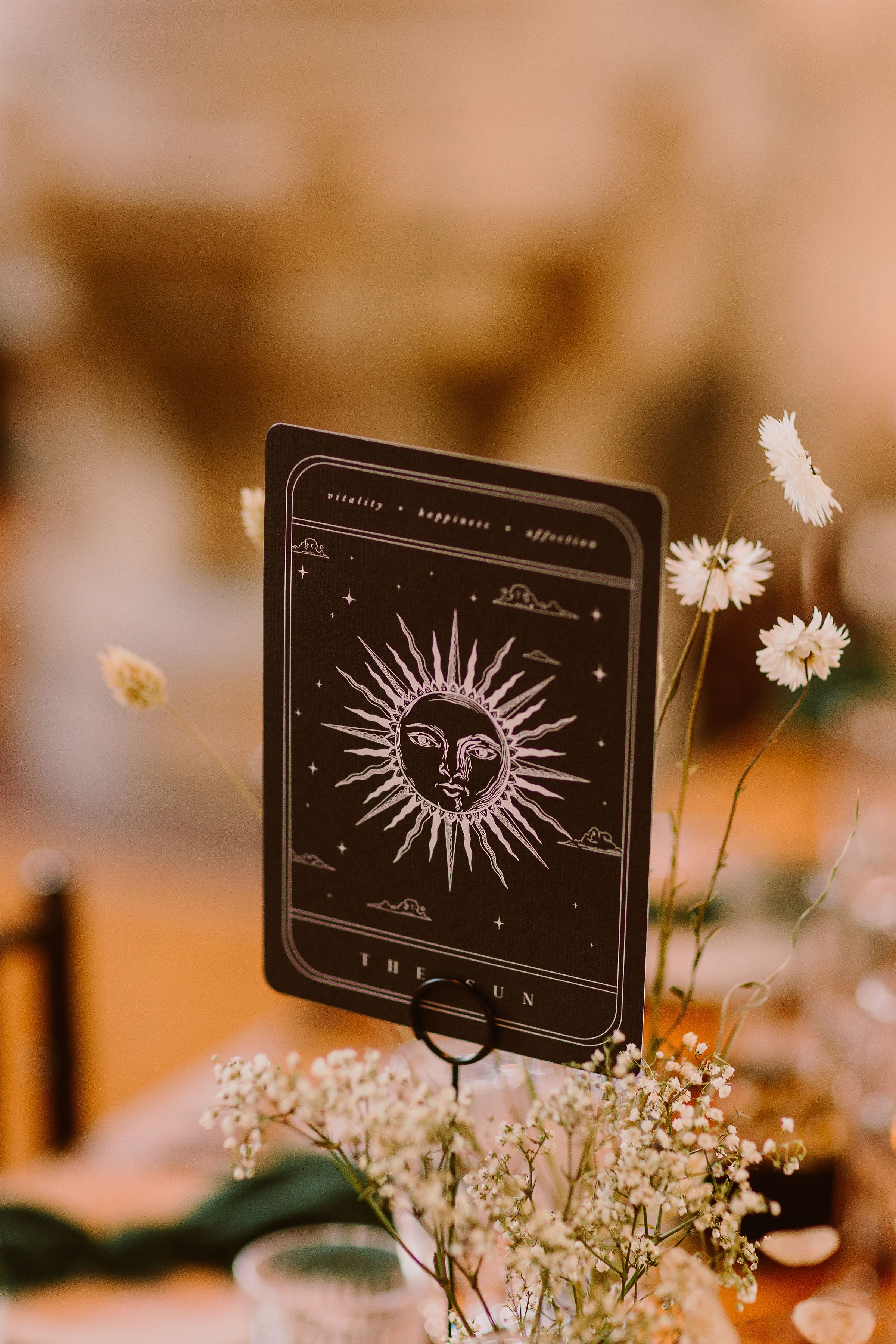 A luxury tarot-inspired wedding stationery card with a celestial sun design is displayed on a cardholder amid romantic white florals.