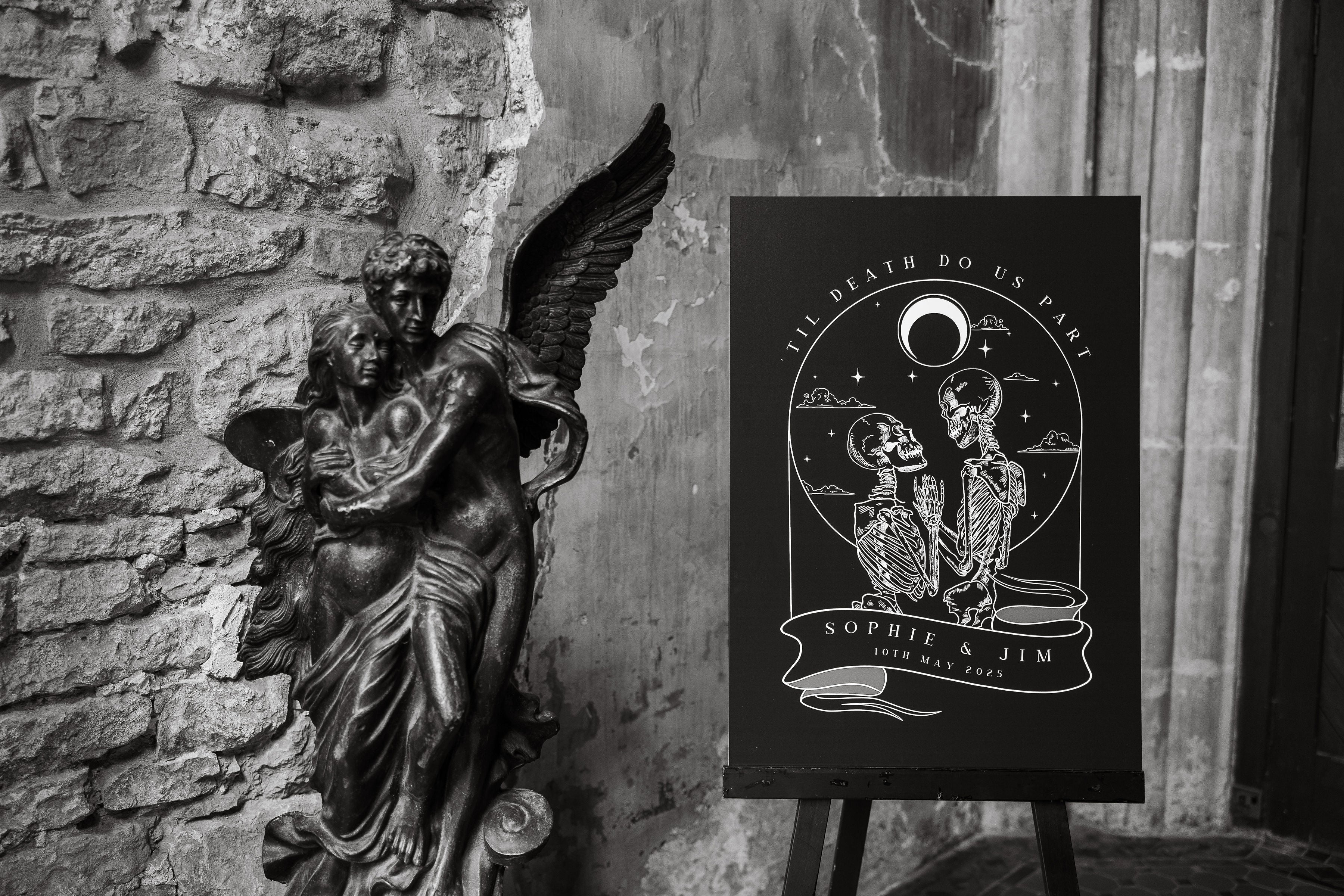 'Til Death Do Us Part Skeleton Lovers wedding welcome board on an easel against a stone wall with an angel statue.