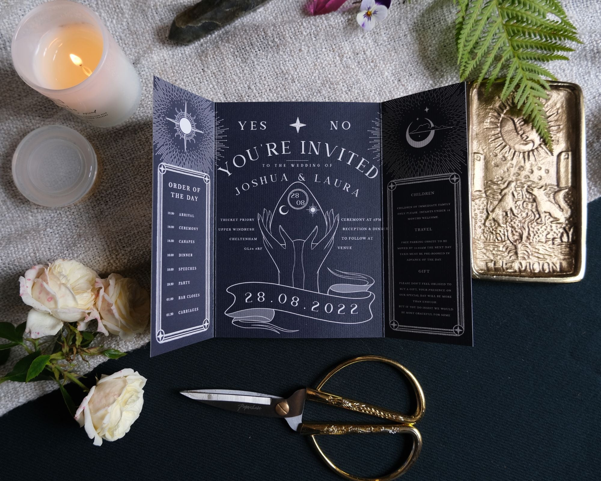 Gatefold wedding invitation in black with celestial design, surrounded by roses, candles, and scissors.