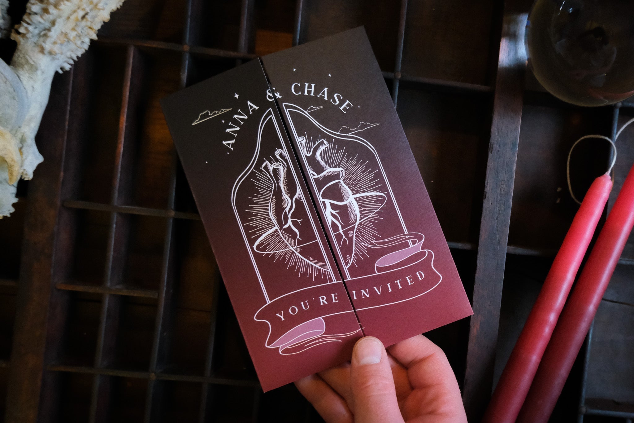 'Bleeding Heart' gatefold wedding invitation displayed on a wooden surface with a hand holding it, featuring customizable colors and a textured matte finish.