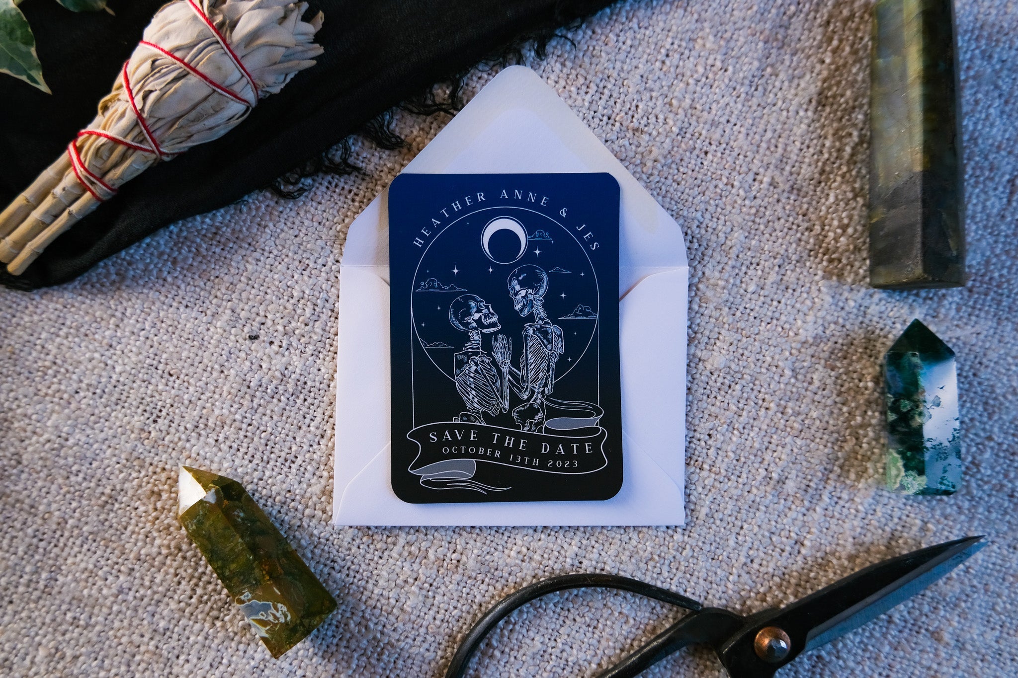 Skeleton Lovers 'Til Death Do Us Part Tarot Save The Date Mini Magnet with white envelope on textured surface surrounded by natural elements.
