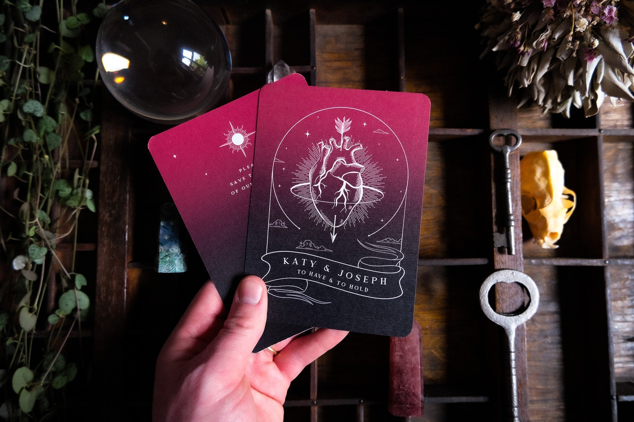 A hand displays a gothic A6 Save the Date card with an anatomical heart, styled on wood with mystical props and dried flowers.