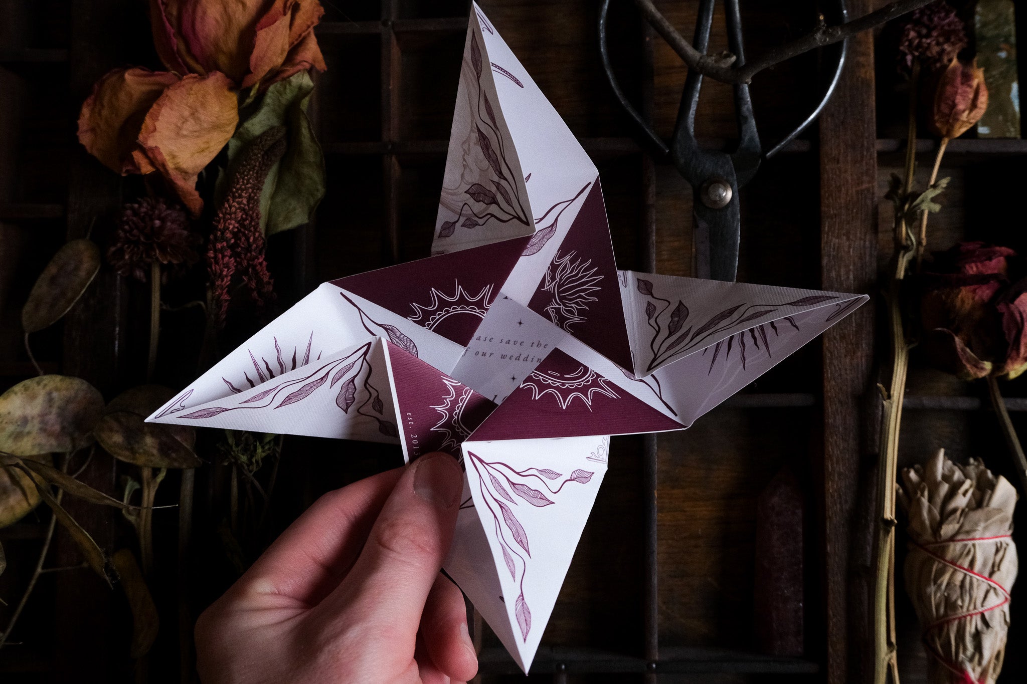 A hand holds a luxury gothic folding wedding stationery piece with celestial maroon and white designs, vintage props on dark wood.