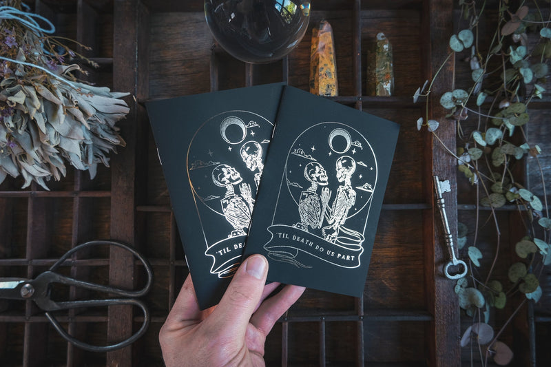 A hand displays two luxury gold foil vow books featuring white skeleton designs; moody gothic backdrop with vintage keys and dried flowers.