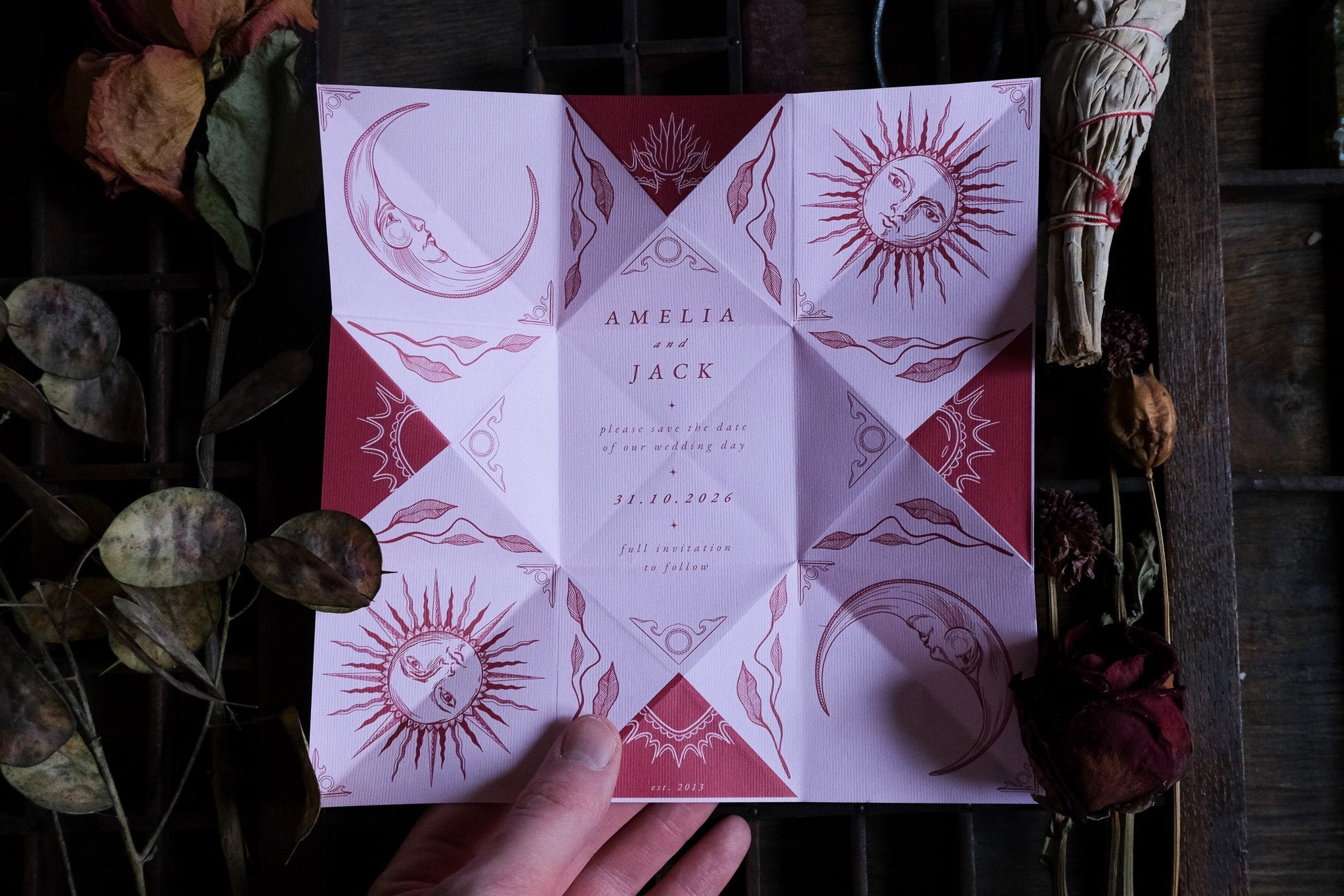 A hand displays a luxury folding letter save the date in deep red and white, with celestial sun and moon motifs, Victorian gothic style.