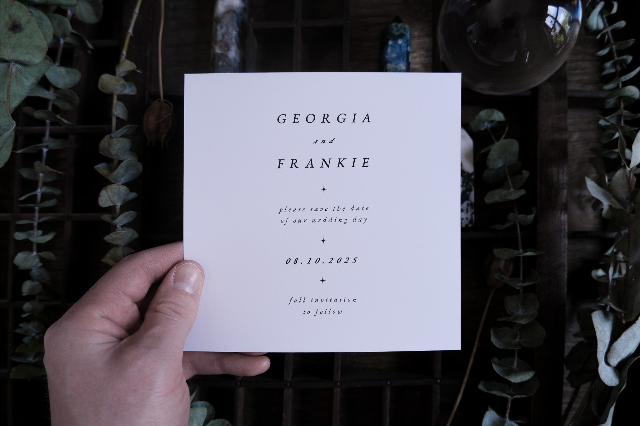 A hand holds a luxury fully foiled wedding save the date card with interlocking rings, set against greenery and elegant decor.