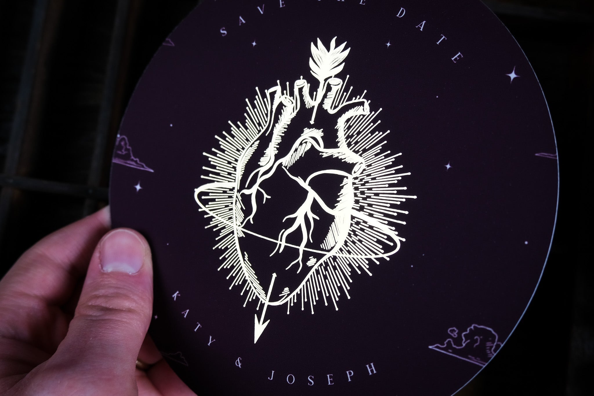 A hand holds a luxury circular foiled save the date card with gothic, romantic design featuring an anatomical heart and radiant detailing.