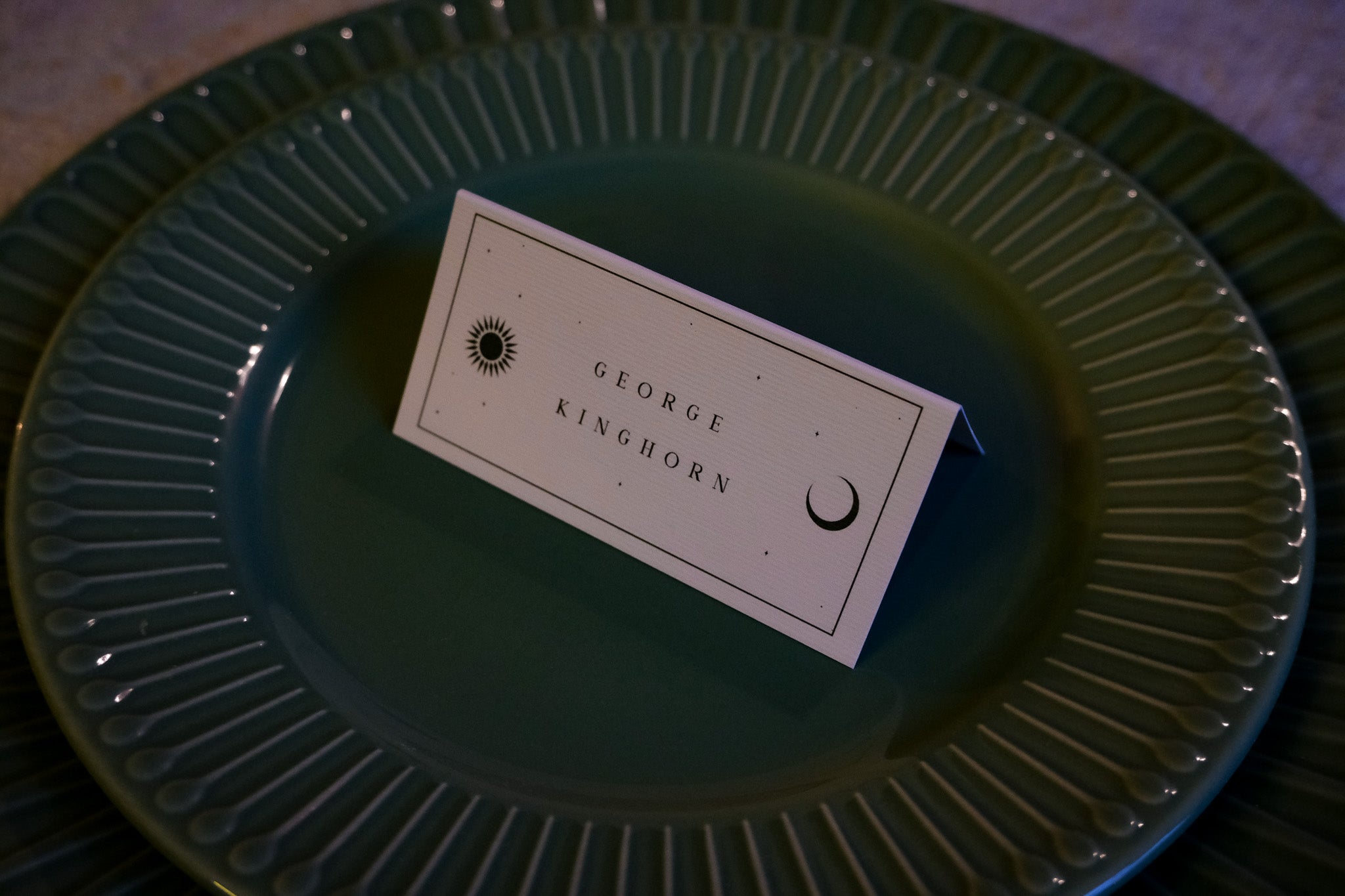 A celestial-themed wedding placecard with sun and moon motifs rests on green ceramic plates, perfect for luxury gothic UK weddings.