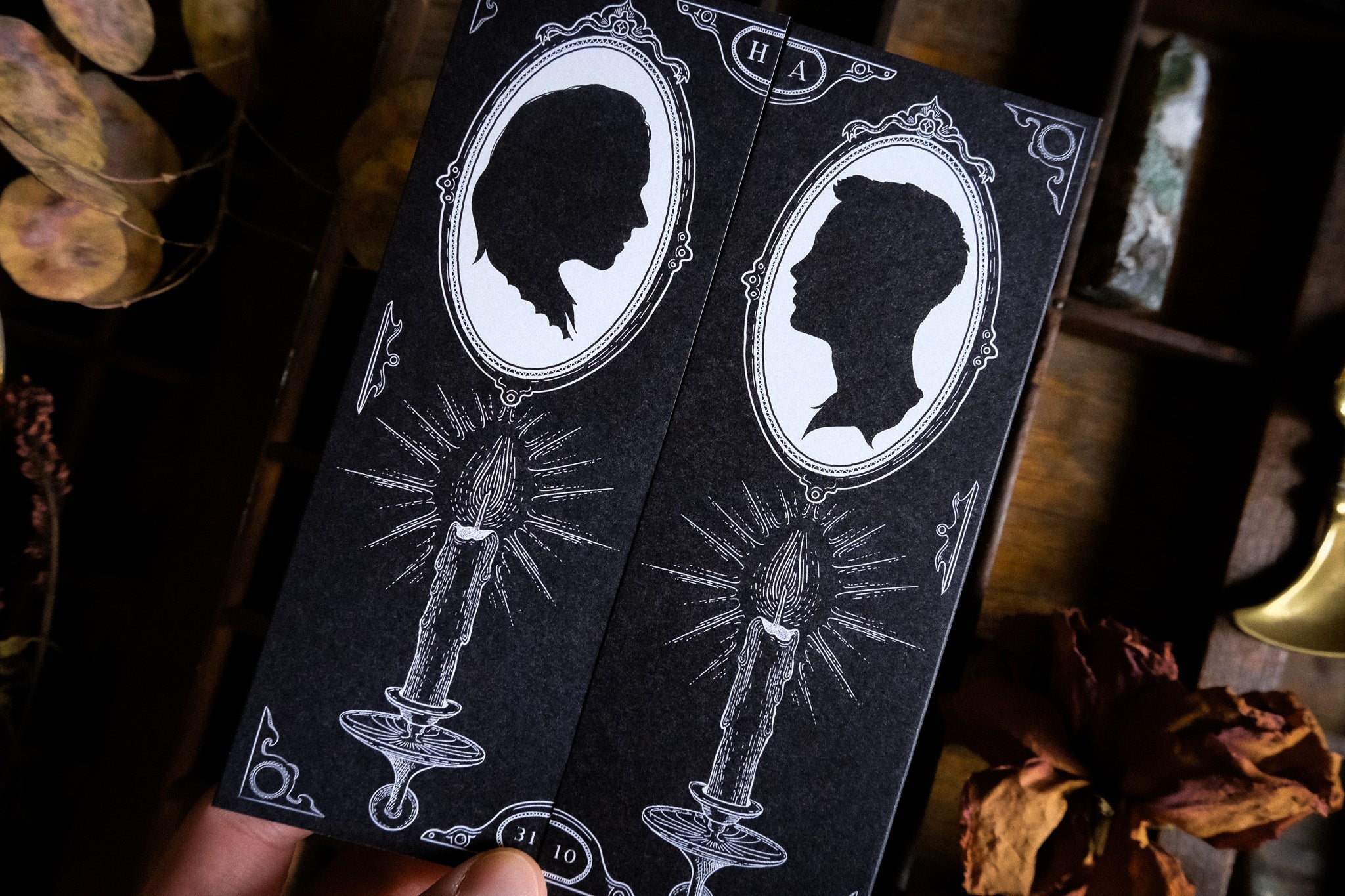 A hand presents two luxurious, gothic gatefold wedding invitations with custom silhouettes and candle motifs, styled amid dried florals.
