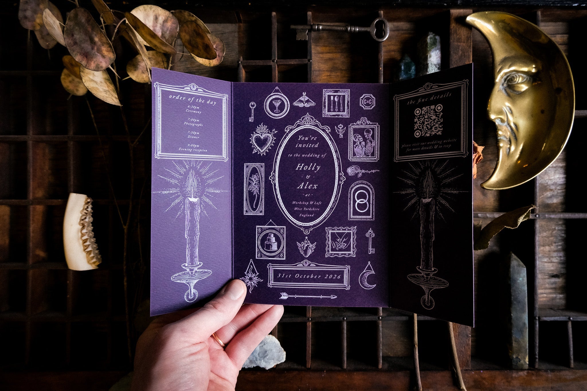 A hand displays a luxury UK wedding gatefold invitation with gothic silhouette design, mystical white symbols, and gold celestial accents.