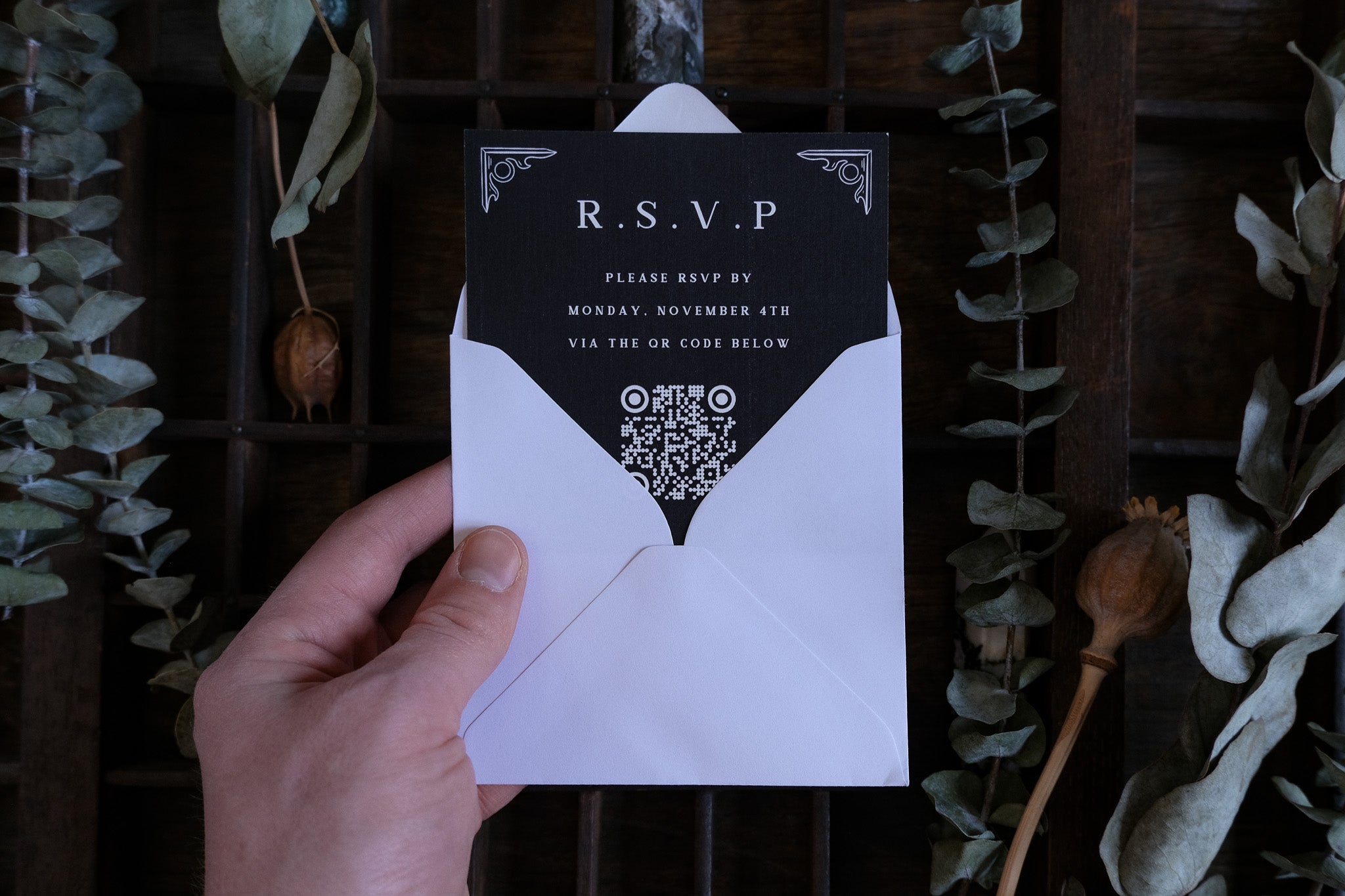 A hand holds a luxury romantic wedding RSVP card in a white envelope, surrounded by dried leaves and rustic wooden decor.