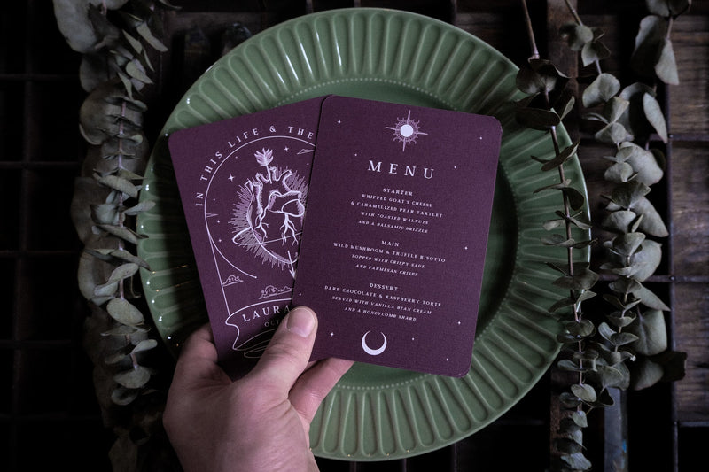 A hand holds two maroon luxury wedding menu cards with white celestial designs in a gothic, romantic style over a green plate, surrounded by dried eucalyptus.