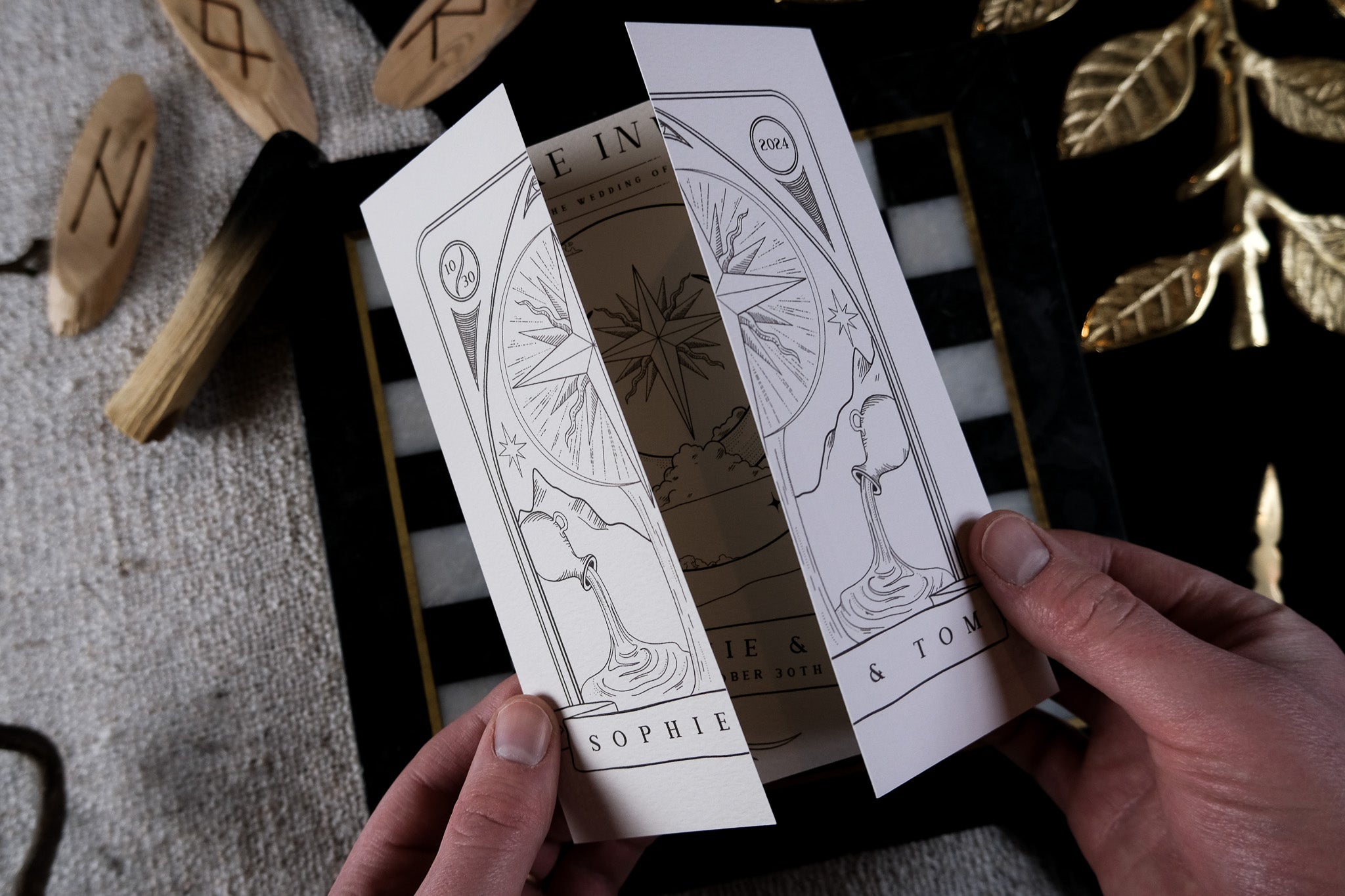 A person holds a luxury gothic gatefold wedding invitation with mystical celestial designs, set among golden leaves and rune tokens.