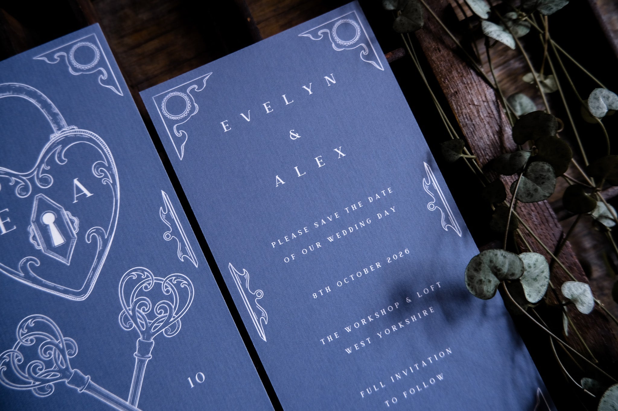 A close-up of a luxury UK save the date card with vintage white motifs on deep blue paper; dried leaves add romantic, dramatic flair.