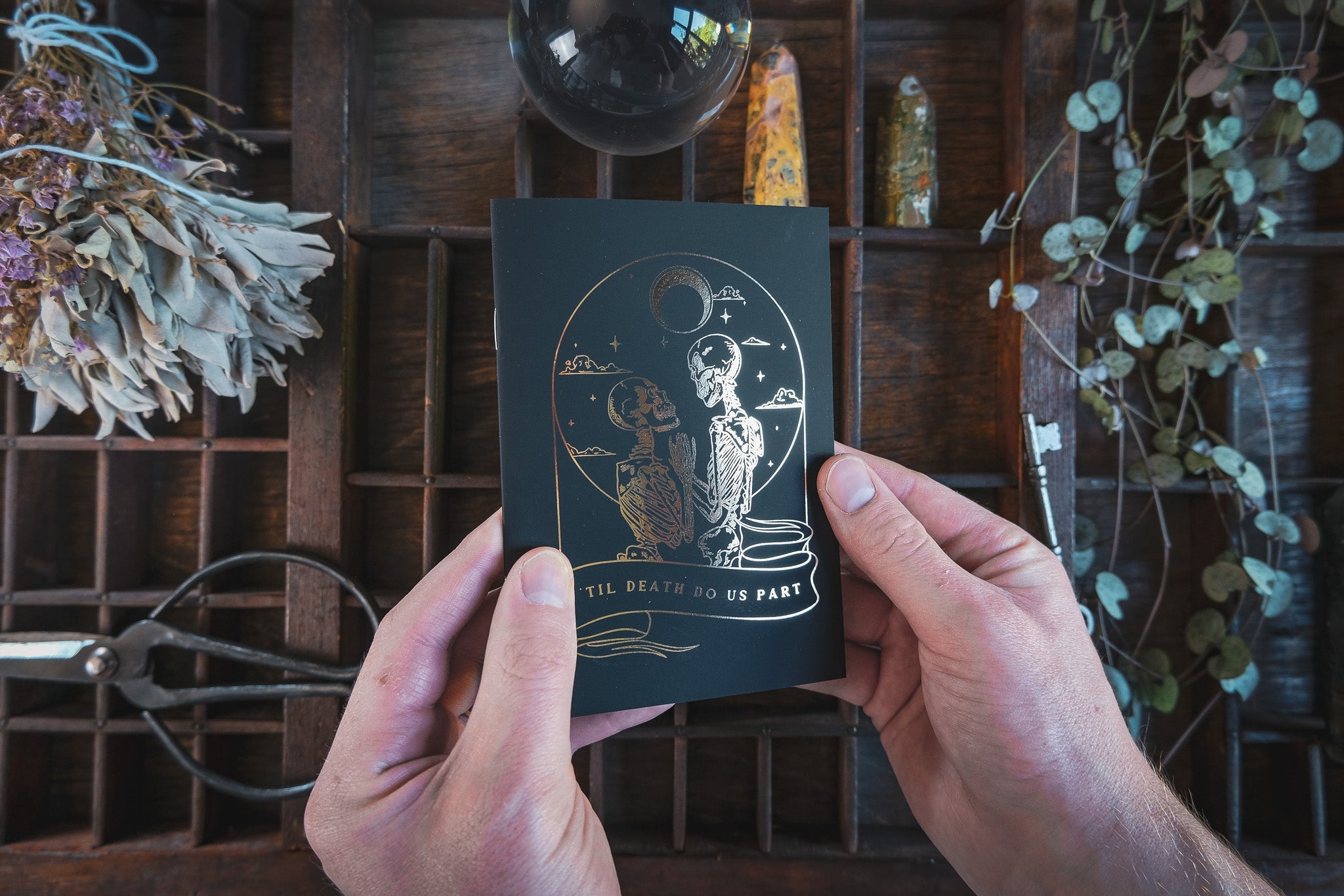 A person holds a black luxury vow book with dramatic gold foil celestial skeleton art, set on wood with dried flowers and art supplies.