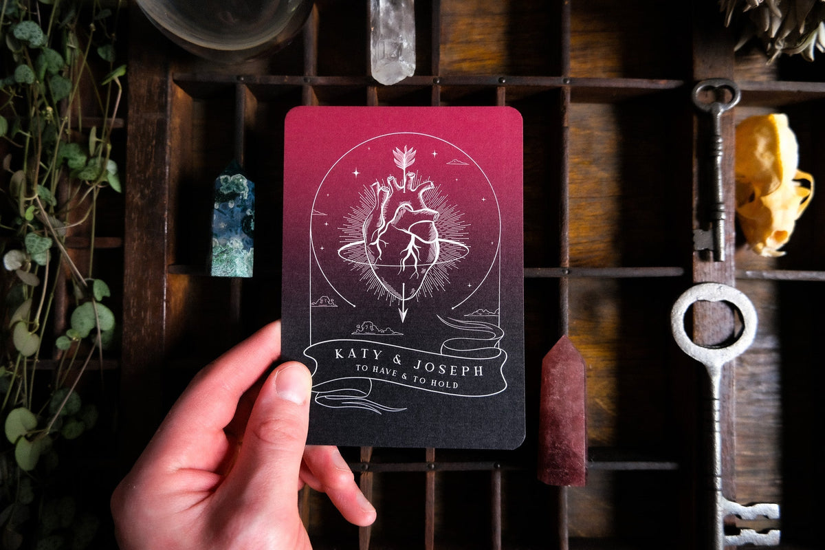 A hand presents a luxury gothic A6 save the date card with an anatomical heart and celestial rays, surrounded by vintage treasures.