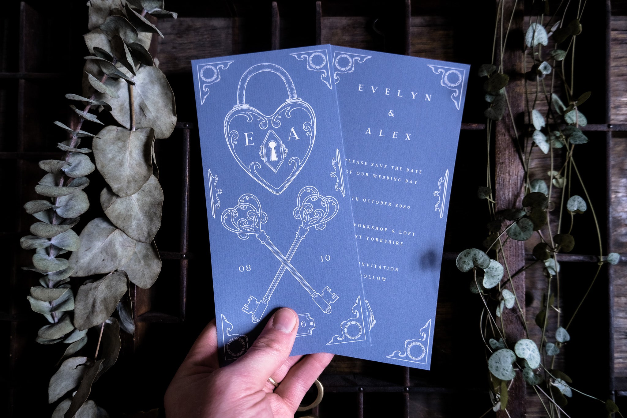 A hand displays two luxury vintage-inspired blue save the date cards with ornate white key and padlock motifs, styled on dark wood with dried greenery.