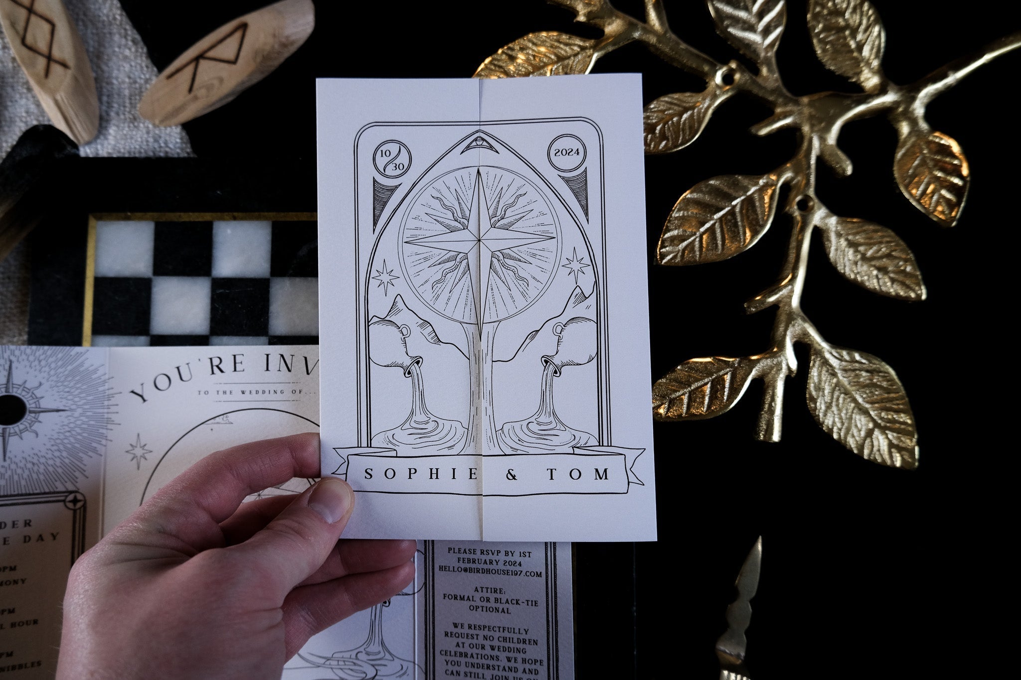 A hand holds a luxury celestial tarot-inspired gatefold wedding invitation with abstract, mystical design on premium card; gothic stationery accents lie behind.