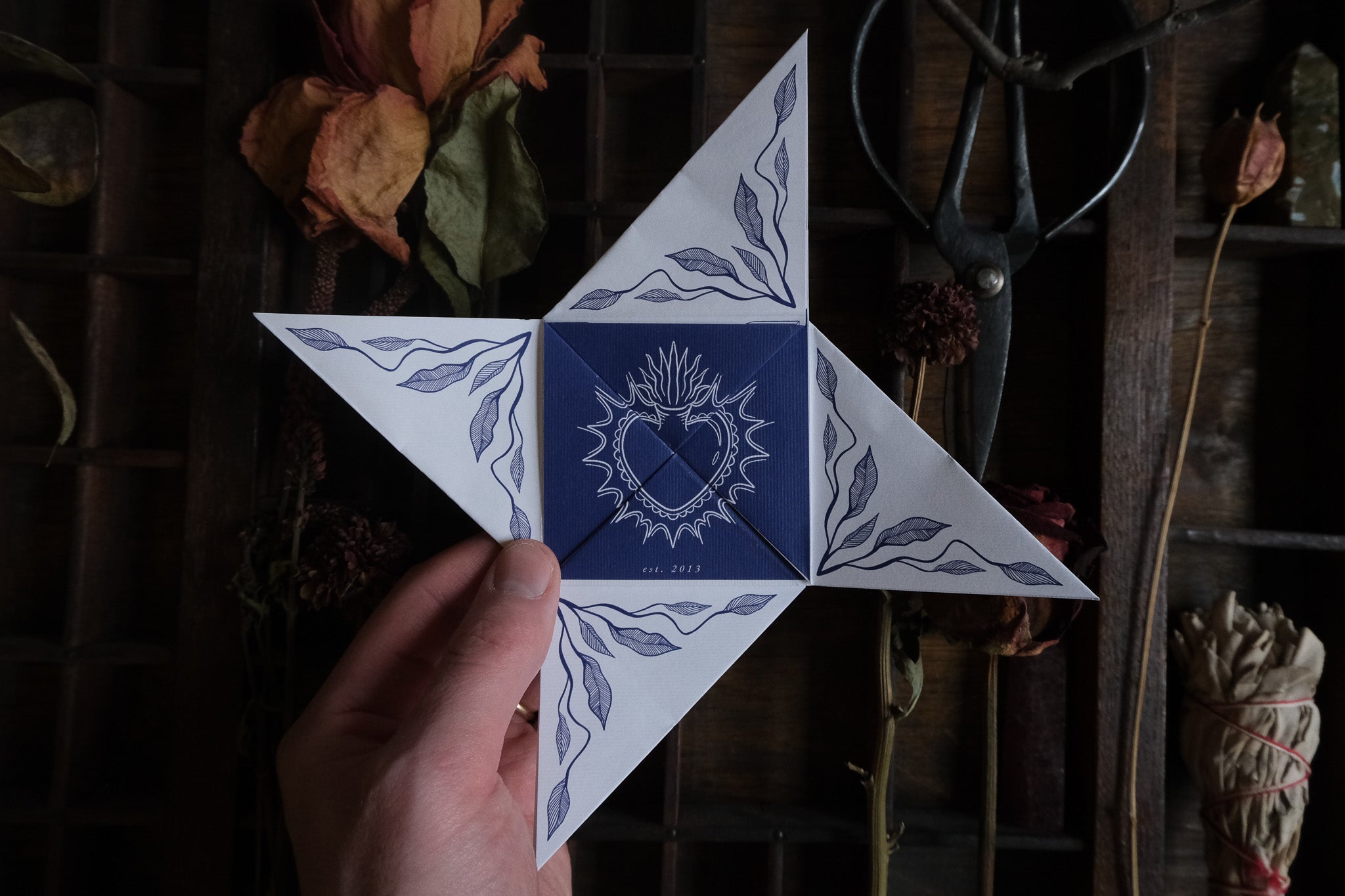 A hand holds luxury celestial self-fold wedding stationery with blue leaf motifs and heart emblem, styled with dried flowers and vintage scissors.