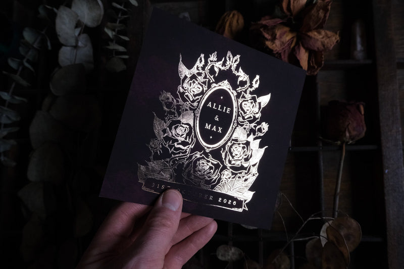 A hand holds a luxury foiled save the date card with dark florals in gothic Victorian style, accented by dried flowers for a dramatic touch.
