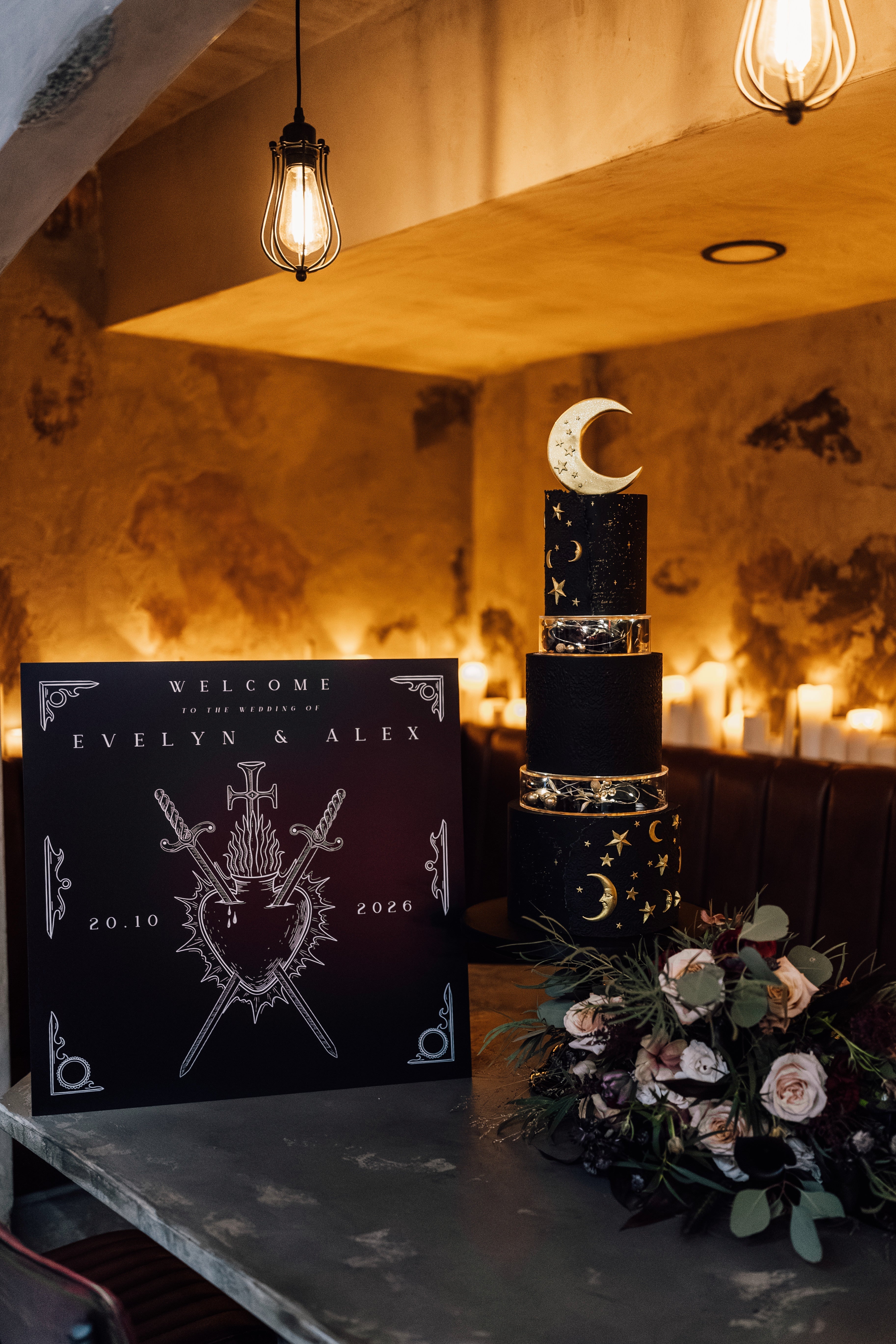 A dramatic, gothic welcome board with celestial foil accents stands beside a black starry cake and romantic florals on a candlelit table.