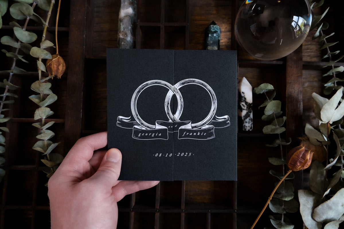 A hand holds a luxury black gatefold wedding invitation with gold foil, set among dried foliage and crystals for a mystical, romantic look.