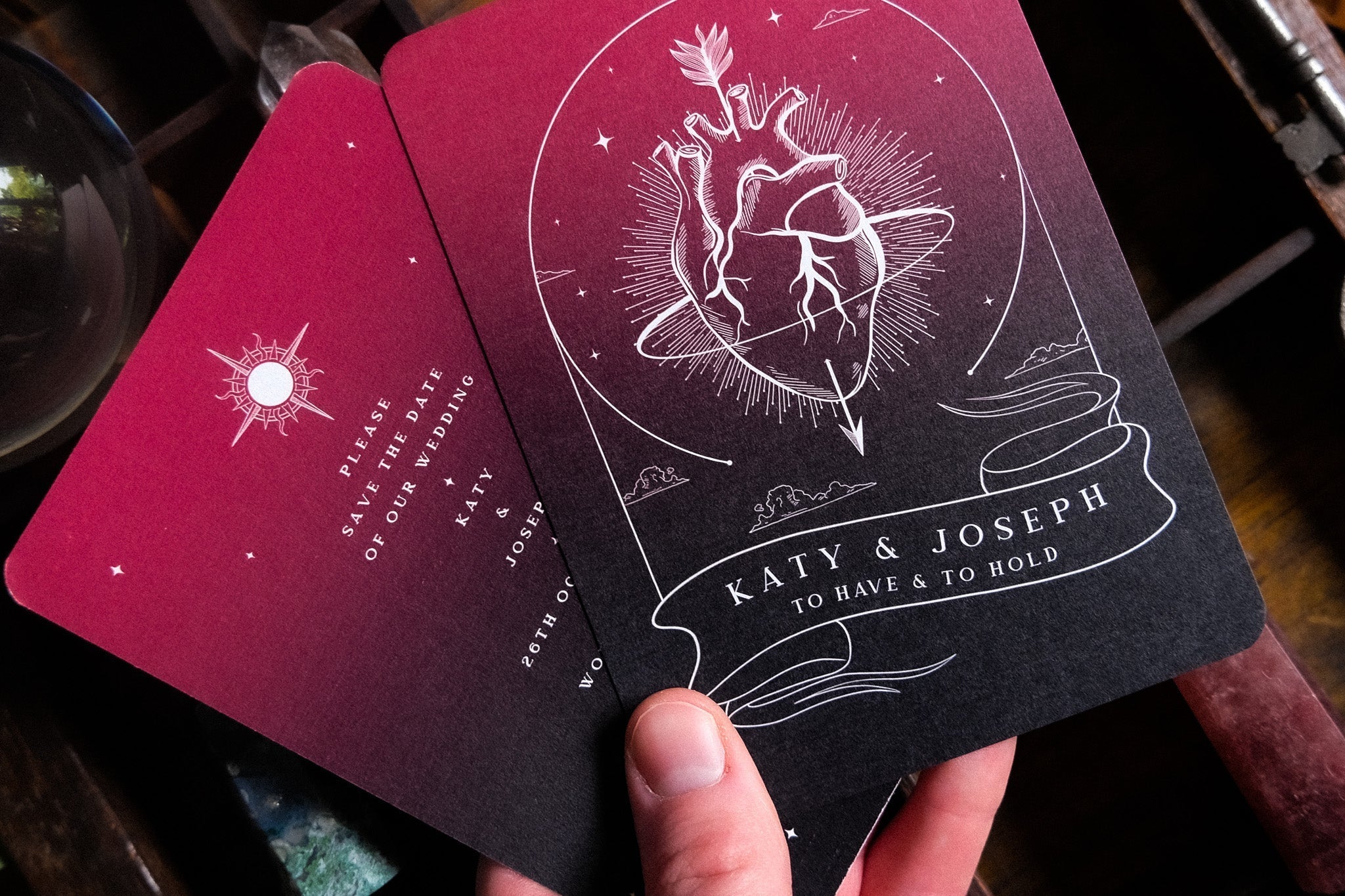A hand displays two luxury A6 gothic save the date cards featuring an anatomical heart tarot motif and dramatic celestial rays.