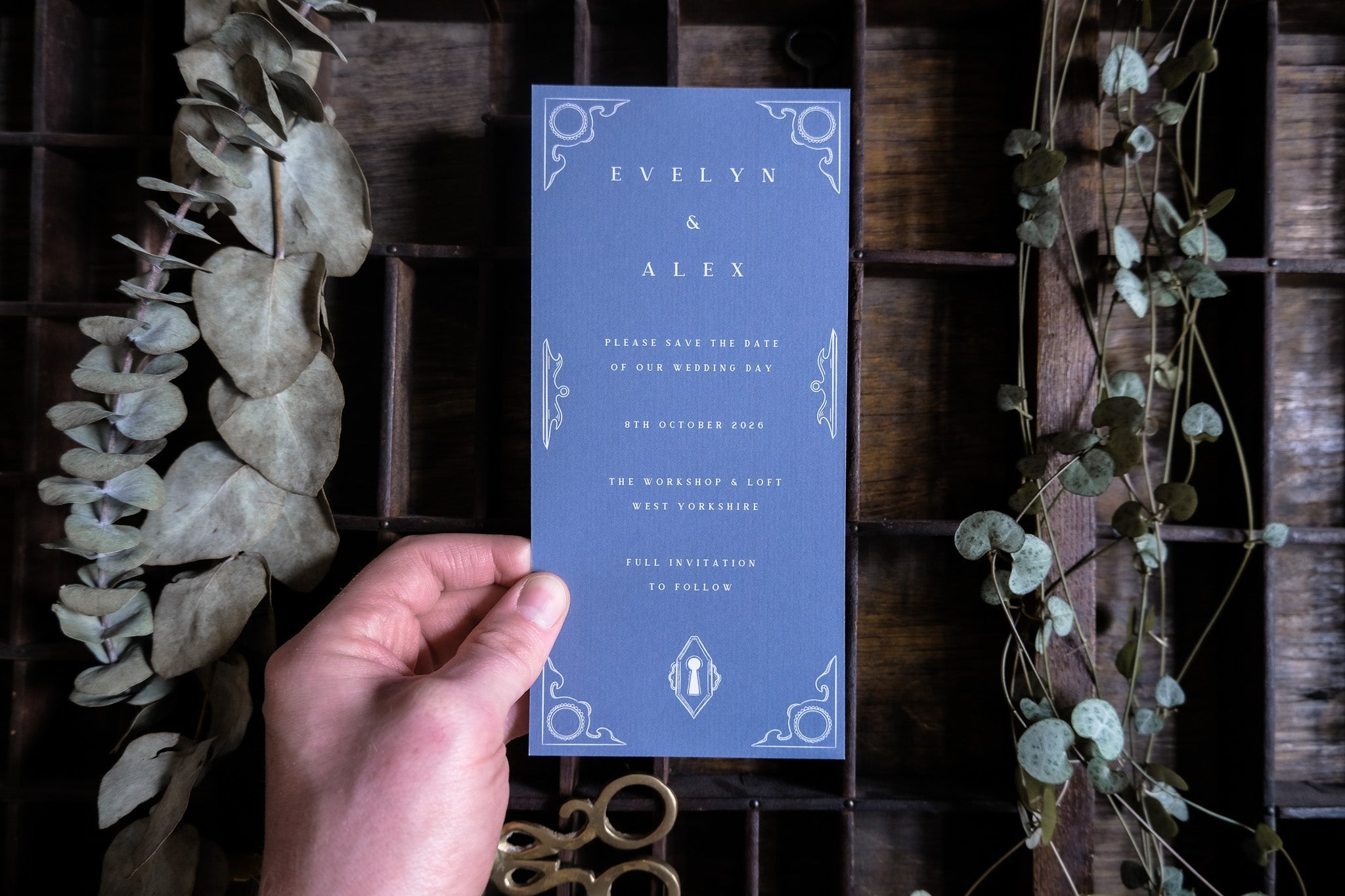 A hand holds a luxury vintage-style wedding invitation with ornate white borders, set among dried leaves on a dark wood table.