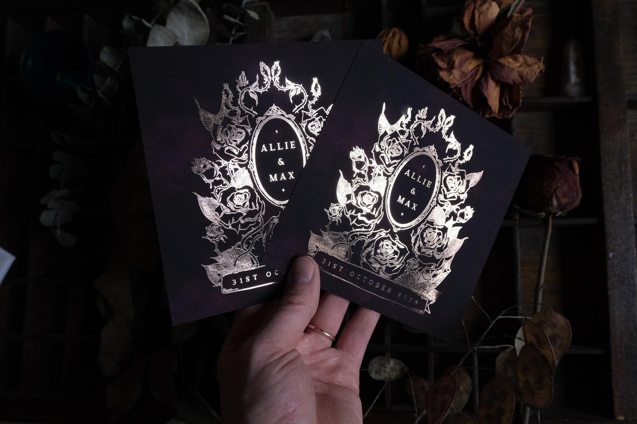 A hand displays two luxury foiled gothic wedding stationery cards with dramatic dark florals and Victorian-inspired design; dried flowers rest behind.
