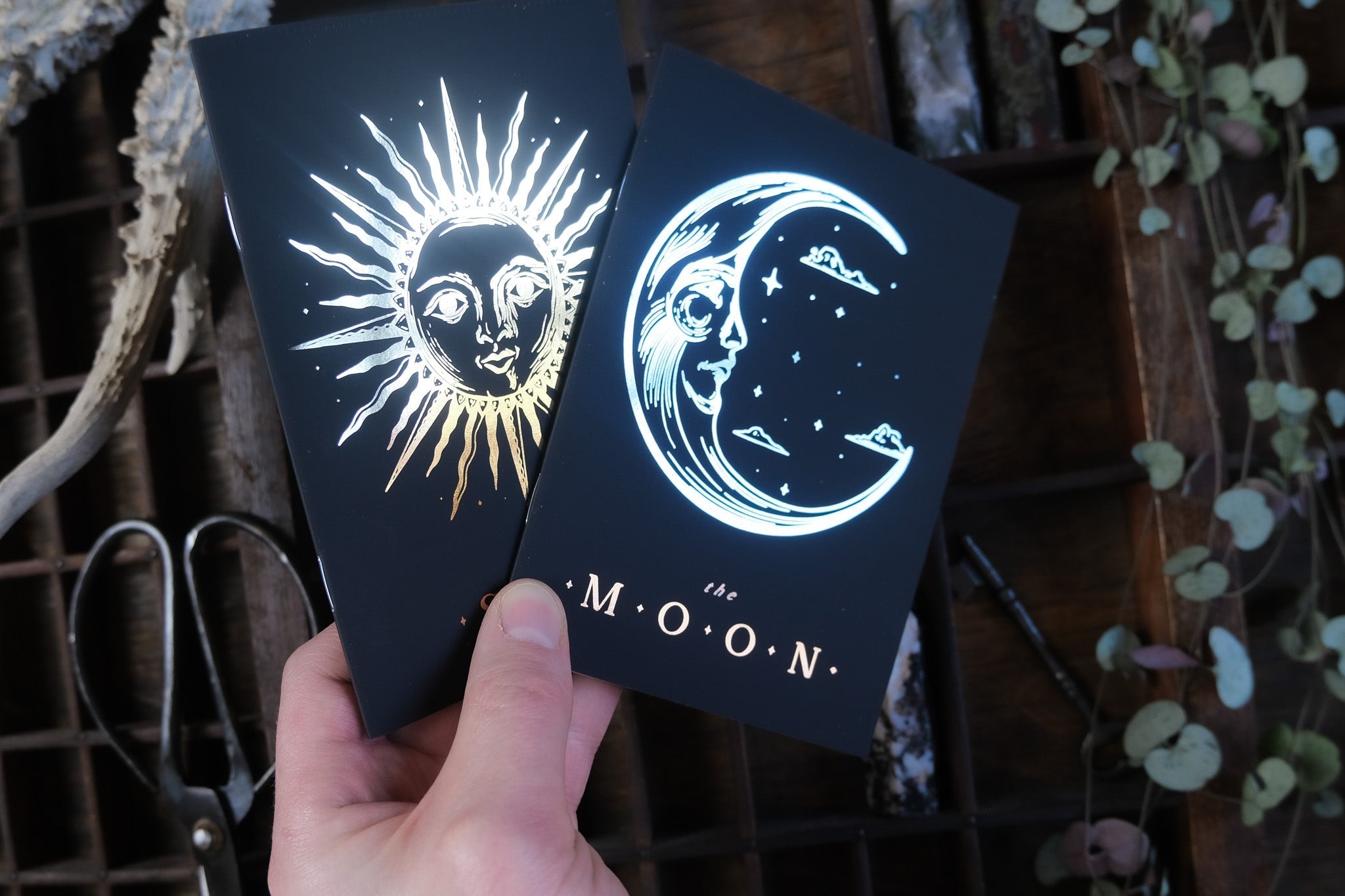 A hand displays luxury gold and silver foiled vow books with celestial sun and moon motifs, set against rustic boho wedding decor.