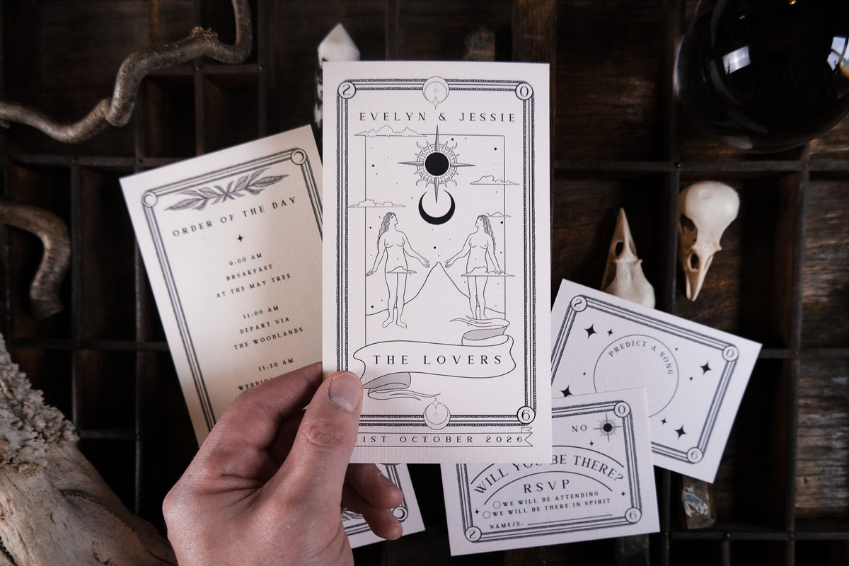 A hand shows luxury gothic tarot-inspired wedding stationery suite with dramatic vintage designs on rustic wood, bones adding a mystical touch.