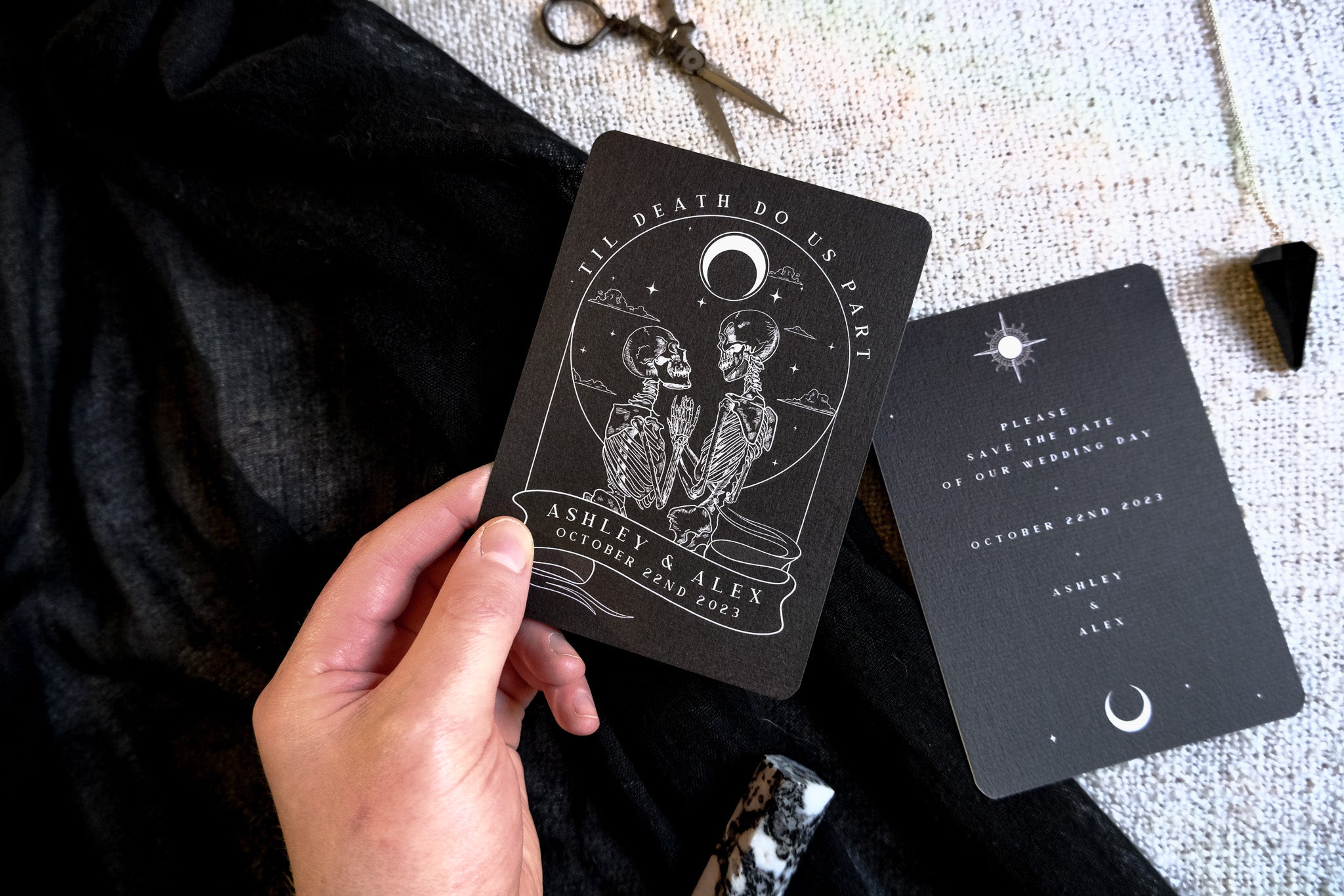 A hand displays luxury gothic wedding stationery with a romantic tarot-inspired design of two skeletons and celestial crescent moon.