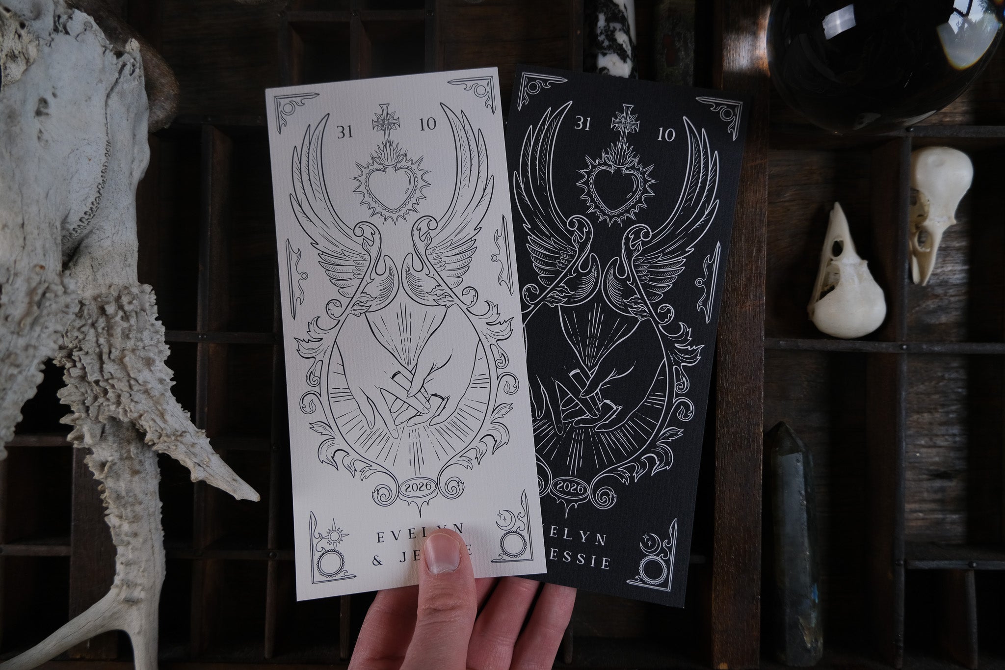 A hand displays two luxury gothic wedding stationery cards, one black, one white, with winged hearts and swords; bones and crystals below.
