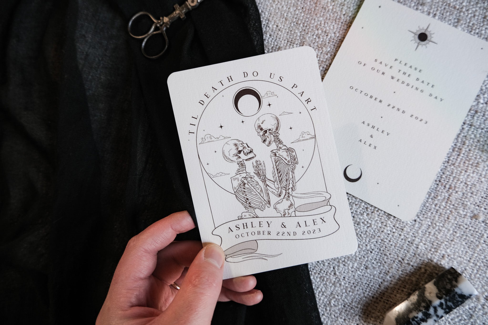 A hand holds a luxury gothic tarot-inspired wedding save the date card with two skeletons and a crescent moon; another celestial card lies nearby.