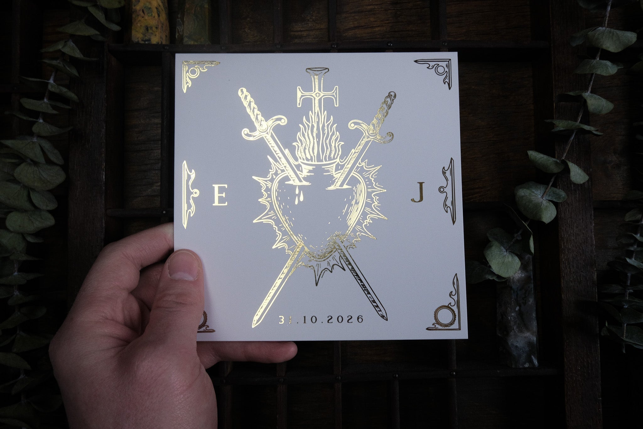 A hand presents a square, luxury gothic save the date card with gold foil embossed mystical heart, sword, and crucifix on crisp white.